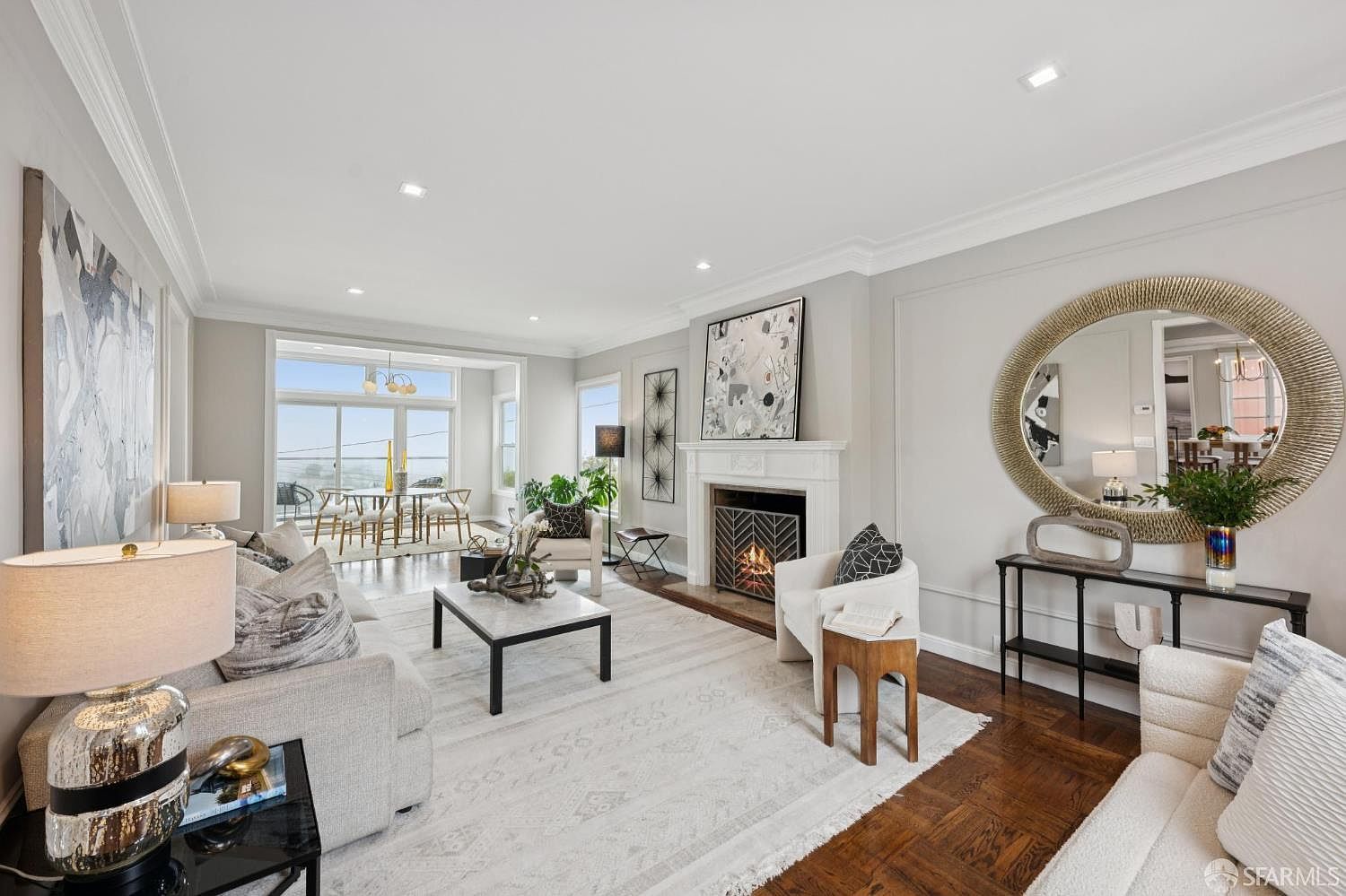 This is a bright and airy living room featuring hardwood floors, a fireplace, and large windows that offer a view. The room is furnished with comfortable seating, including sofas and armchairs, arranged around a coffee table and a decorative rug. A large round mirror and artwork add visual interest to the space, creating an inviting and stylish atmosphere.
