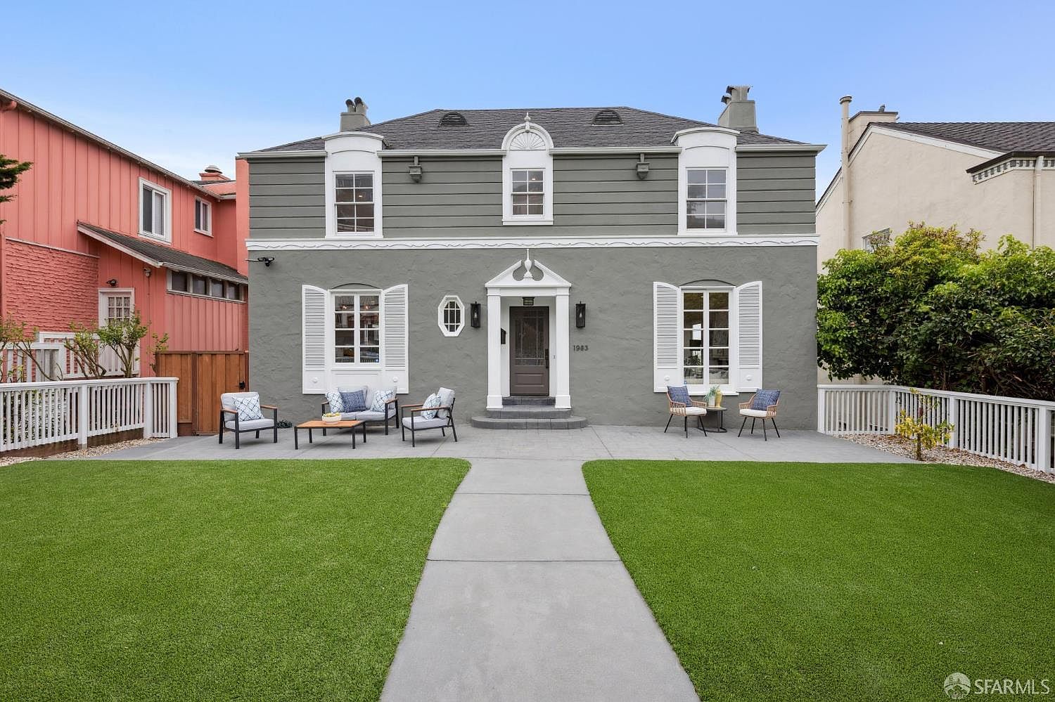 This is a front exterior view of a two-story house with a well-manicured lawn. The house features a gray facade with white trim, symmetrical windows with shutters, and a central entryway with a dark-colored door. Outdoor seating areas are arranged on either side of the walkway, enhancing the curb appeal and inviting atmosphere.