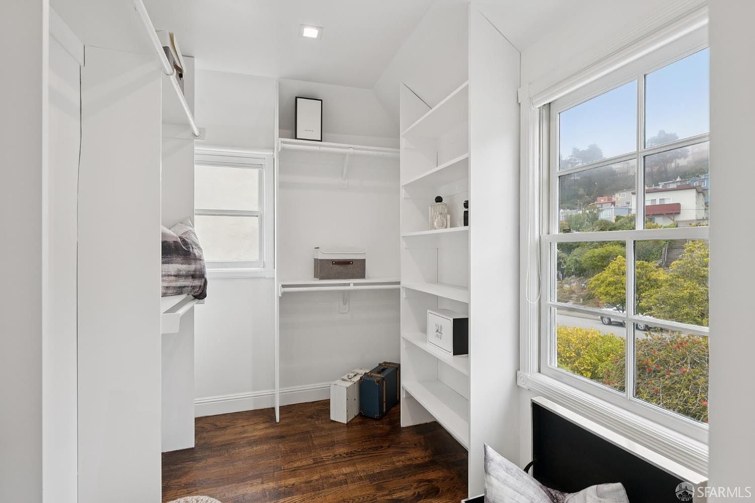 This is an interior shot of a walk-in closet featuring white shelving and dark wood flooring. The closet is well-lit, with a window providing natural light and a view of the outdoors. The space appears organized and functional, offering ample storage.