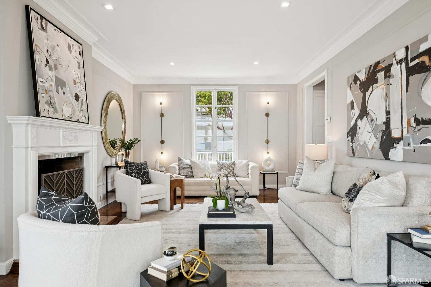 This is an interior shot of a well-lit living room featuring two white sofas, a white armchair, and a fireplace. The room is decorated with modern art, a round mirror, and a black and white patterned rug. The overall impression is elegant and inviting, with a focus on neutral colors and clean lines.