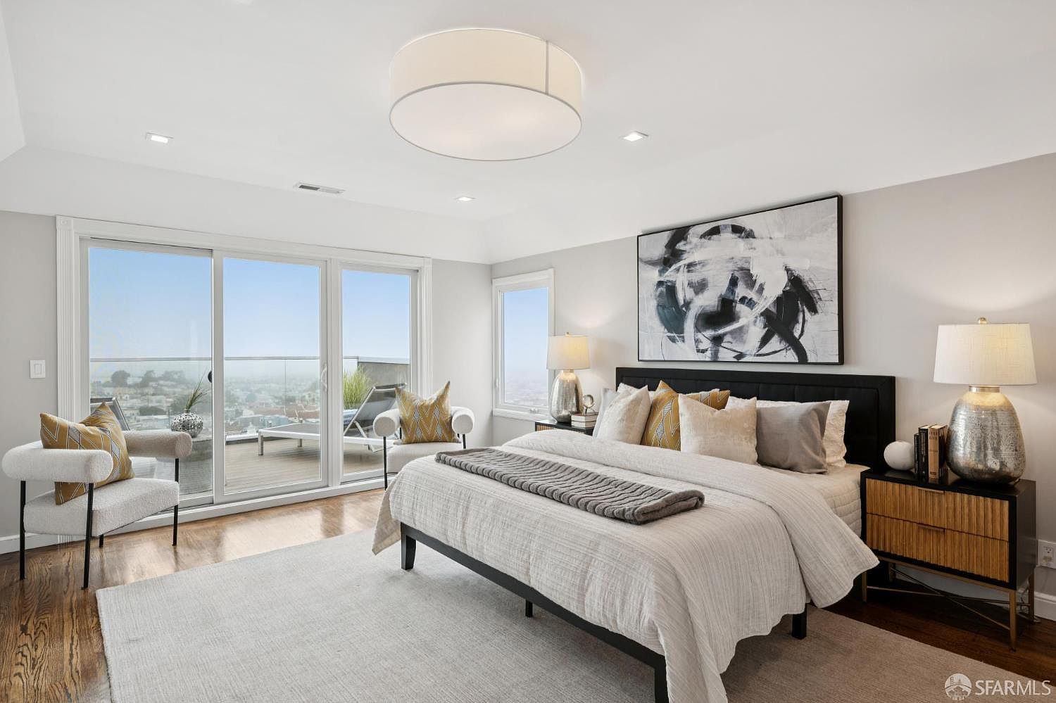 This is a bright and airy primary bedroom featuring a large bed with a black headboard, neutral-toned bedding, and a gray throw. The room has a modern aesthetic with a large abstract art piece, a stylish nightstand with a lamp, and access to a balcony with city views through sliding glass doors. The hardwood floors and light gray walls contribute to the room's serene and upscale ambiance.