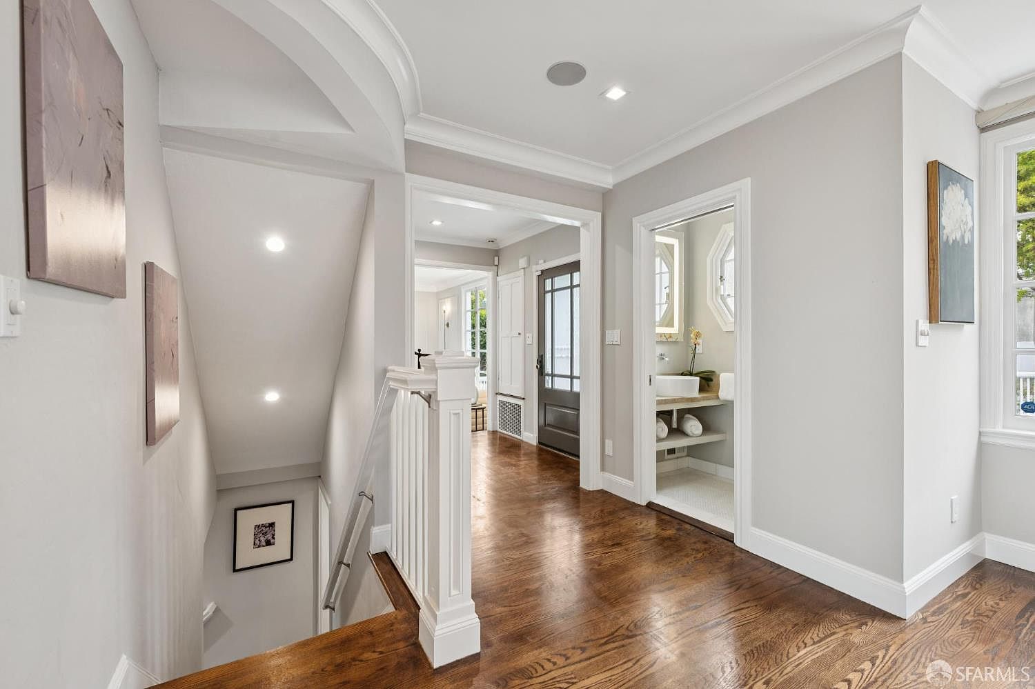 This interior shot showcases a well-lit hallway and staircase. The hallway features hardwood floors, white trim, and neutral-colored walls, leading to a powder room and other areas of the home. The staircase has a white banister and leads to the lower level, creating a bright and inviting transition between floors.