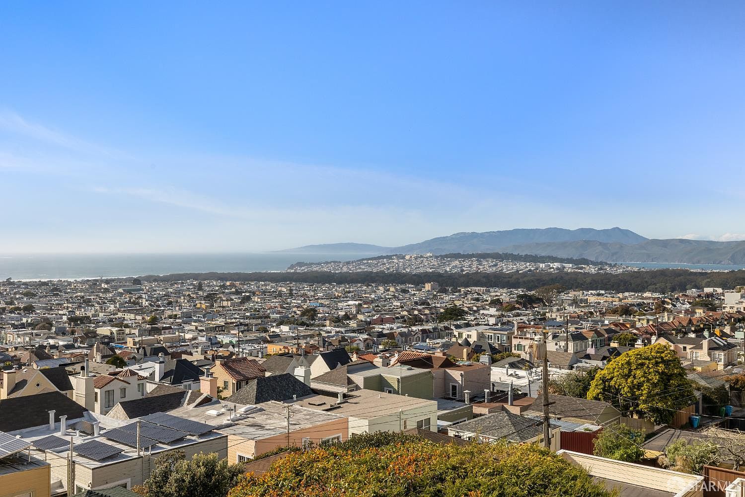 This aerial view showcases a densely populated residential area with a clear blue sky above. The landscape includes a distant coastline and mountains, providing a scenic backdrop to the urban sprawl. The perspective offers a comprehensive overview of the neighborhood's layout and its proximity to natural features, highlighting the location's appeal.