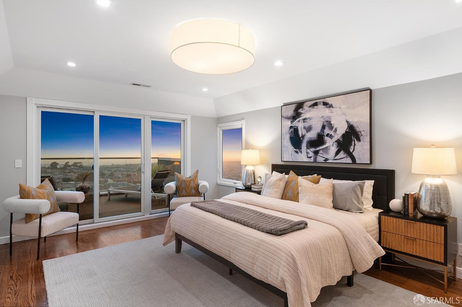This is a bright and airy primary bedroom featuring a large bed with a neutral-toned comforter and decorative pillows. The room has a modern aesthetic with a large abstract art piece above the bed and a stylish nightstand with a lamp. Sliding glass doors lead to a balcony with a view, enhancing the room's appeal.