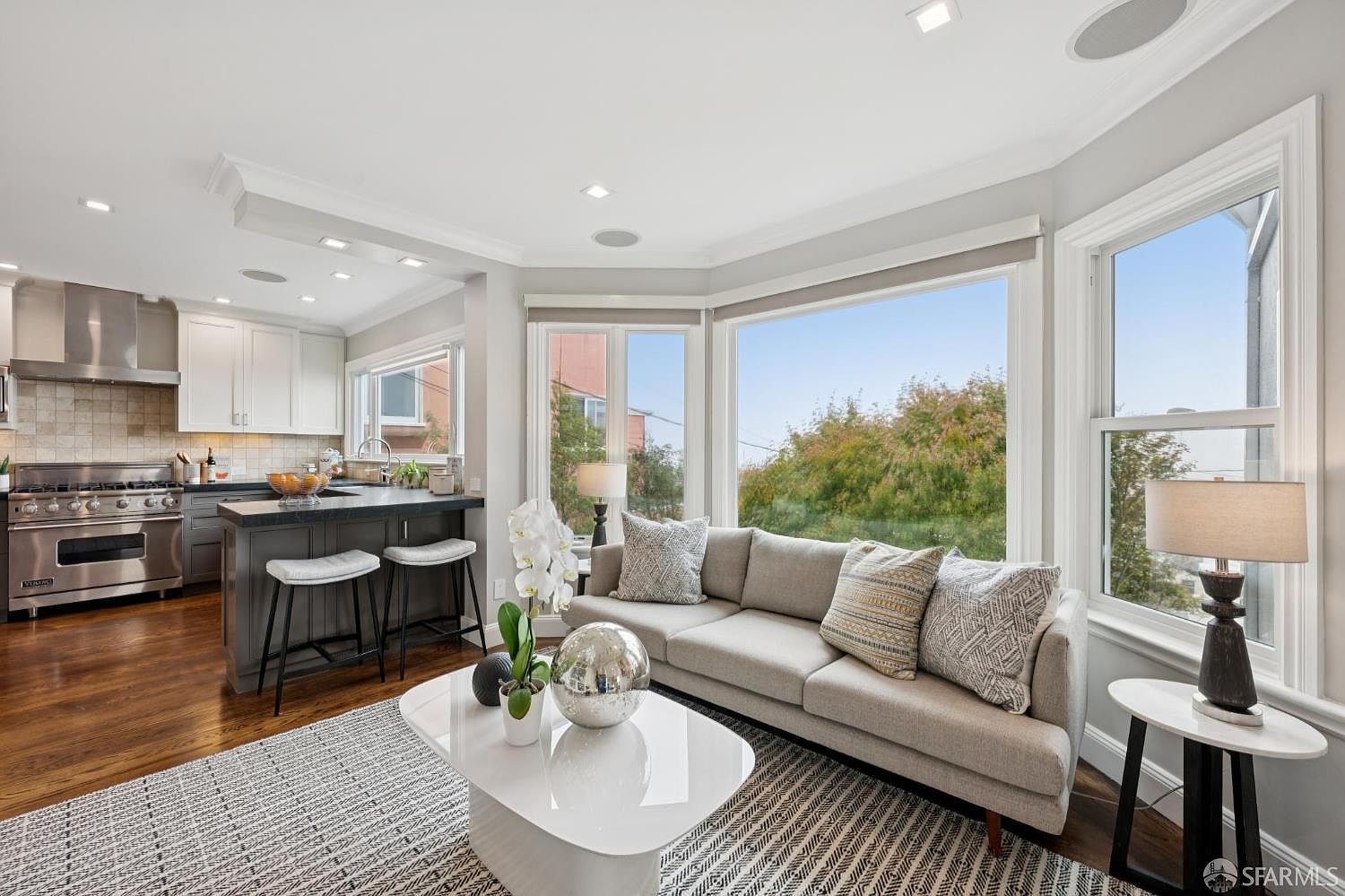 This is an interior shot of a living room that seamlessly blends into a modern kitchen. The living room features a comfortable sofa, stylish pillows, a contemporary coffee table, and a large window offering natural light and a view. The kitchen area includes stainless steel appliances, dark countertops, and bar stool seating, creating an open and inviting space.