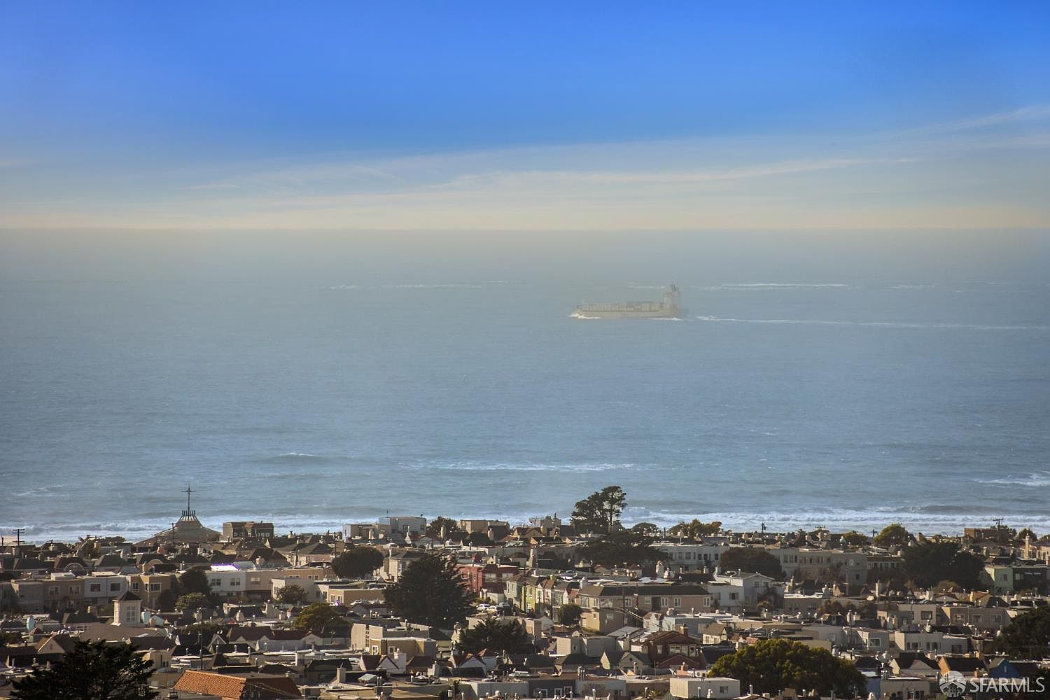 This aerial view showcases a dense residential neighborhood stretching towards the ocean. The houses feature a variety of architectural styles and colors, creating a visually interesting landscape. In the distance, a large cargo ship sails across the water under a clear blue sky, adding a sense of scale and tranquility to the scene.