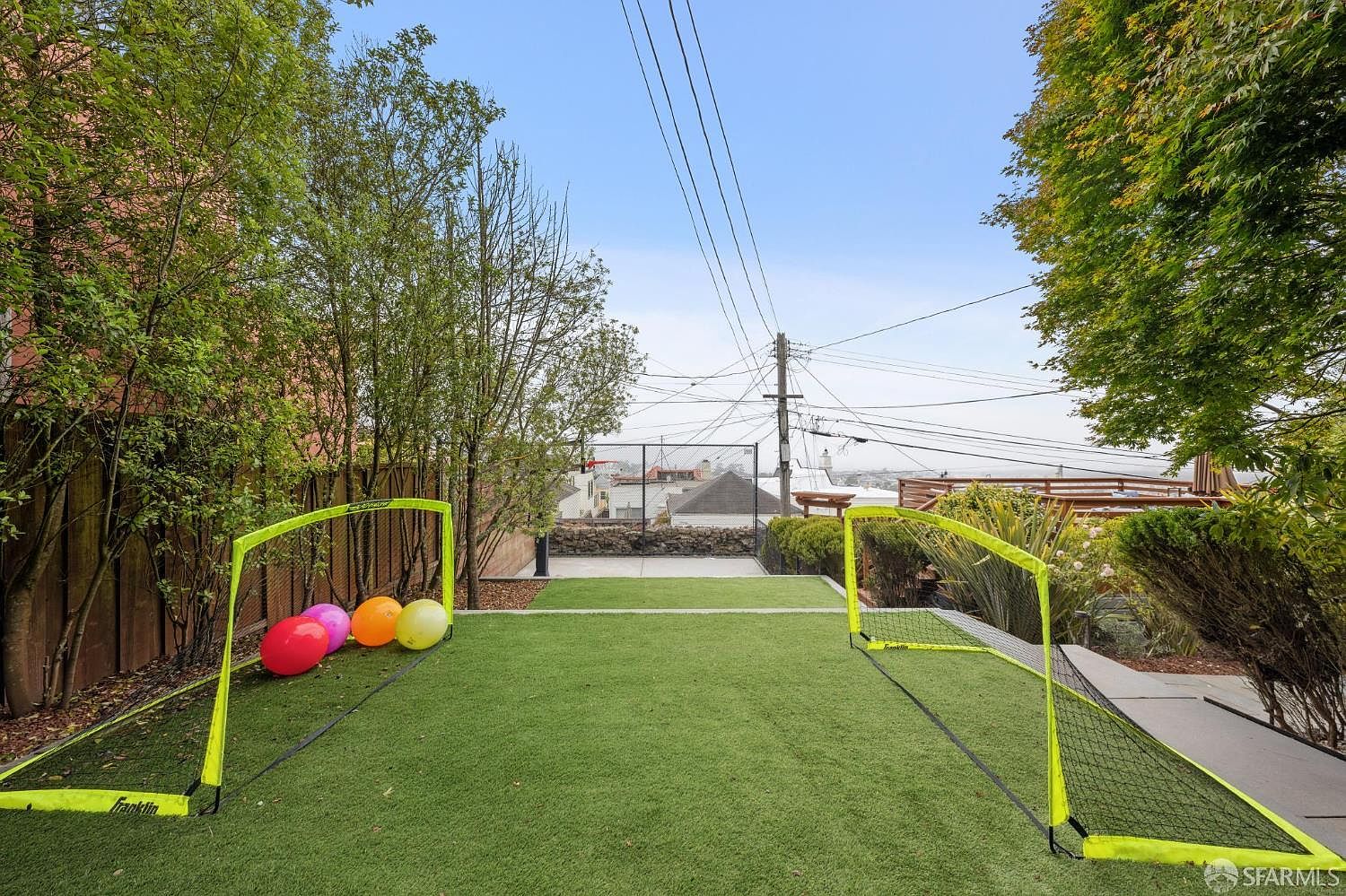 This image showcases a well-maintained backyard featuring artificial turf, two soccer goals, and colorful balls. The yard is bordered by trees and a wooden fence, providing privacy. In the background, there's a glimpse of the neighborhood, creating a sense of community.