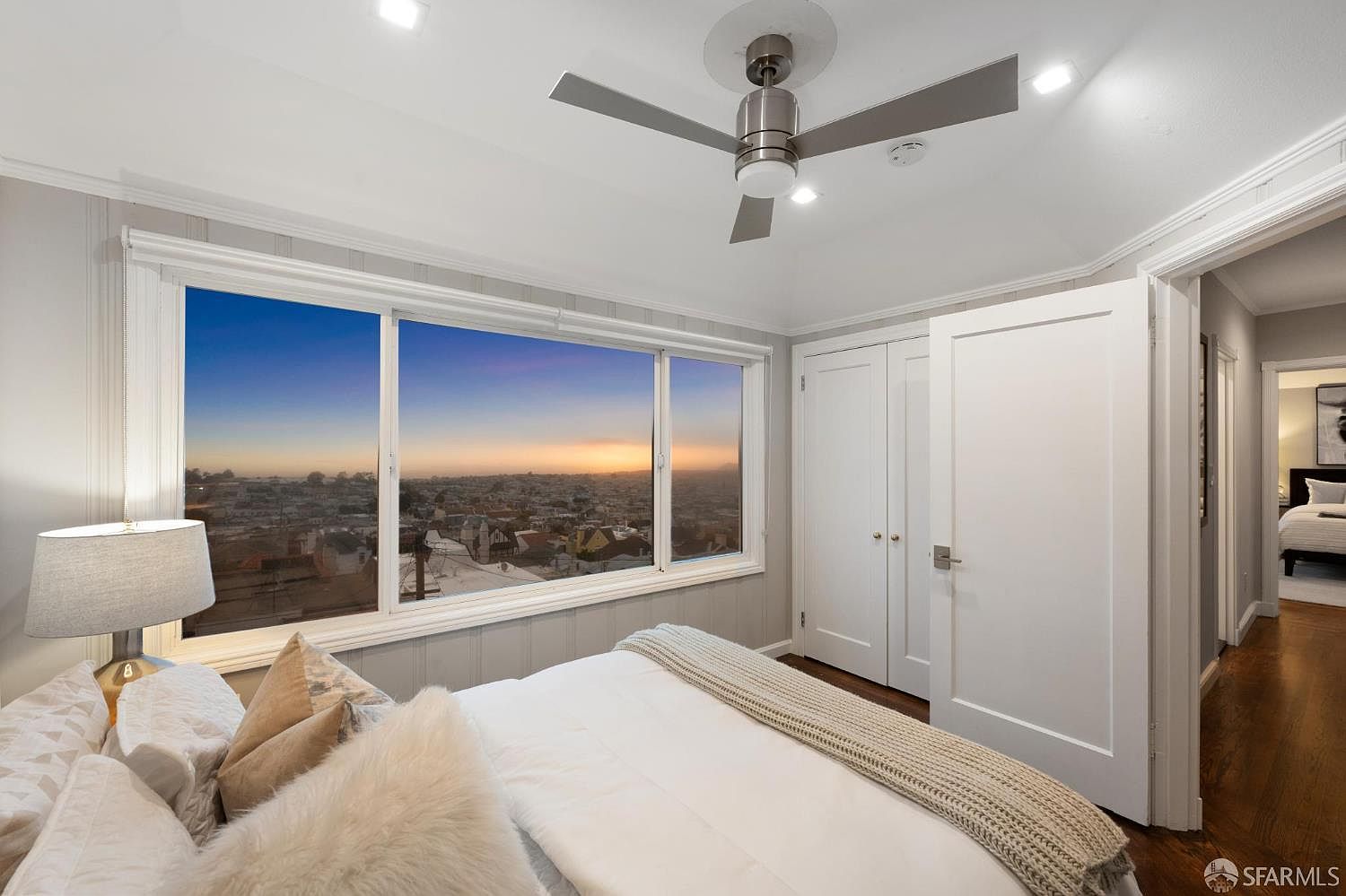 This is a bright and airy primary bedroom featuring a large window with a cityscape view at sunset. The room is decorated in neutral tones with white bedding, light wood accents, and a ceiling fan. A closet and doorway lead to other parts of the home, showcasing the bedroom's integration within the house.