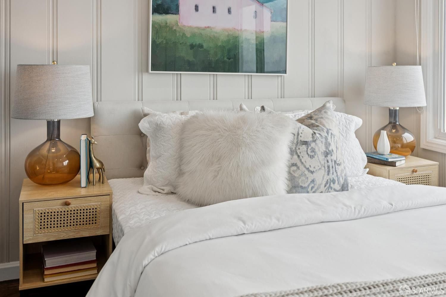 This is a cozy primary bedroom featuring a neutral color palette. The bed is adorned with plush pillows and a white duvet, complemented by bedside tables with lamps. An artwork hangs above the bed, adding a touch of color to the serene space.