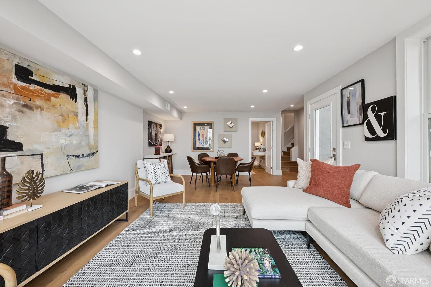 This is an interior shot of a living room featuring a modern design with hardwood floors and a neutral color palette. The room includes a large sectional sofa with decorative pillows, a black coffee table, and a black media console with artwork above. A dining area is visible in the background, creating an open-concept living space.