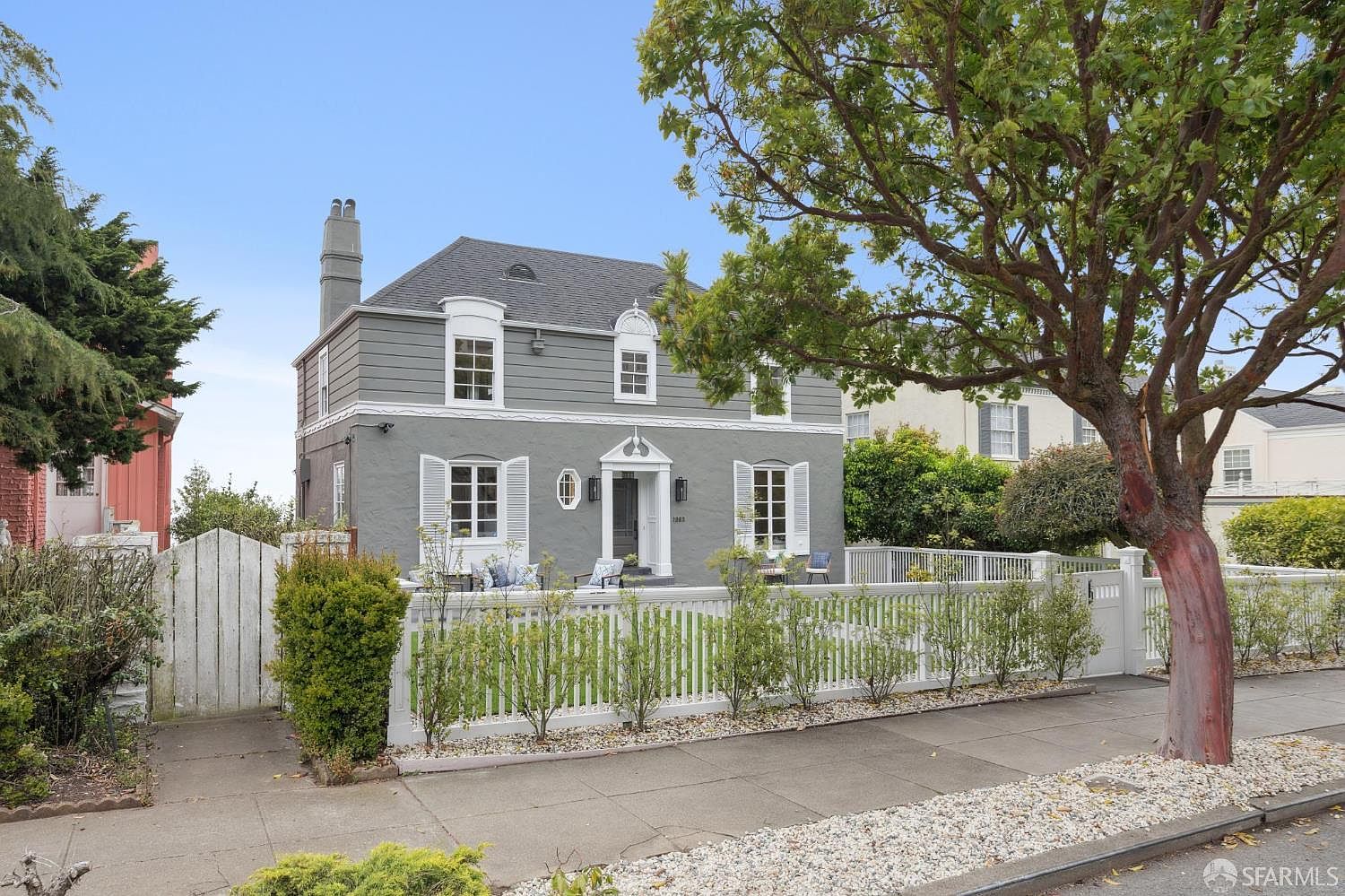 This is a charming two-story house with a gray exterior and white trim. The house features a well-manicured front yard enclosed by a white picket fence, adding to its curb appeal. A mature tree partially frames the house, providing shade and enhancing the overall aesthetic.