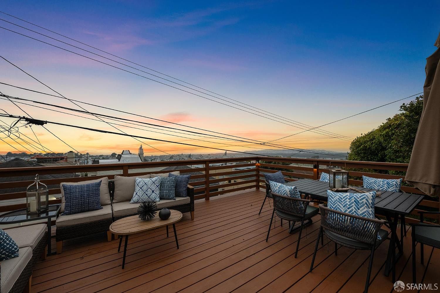 This image showcases a beautifully furnished outdoor deck or balcony, perfect for relaxation and entertaining. The space features comfortable seating arrangements, including a sofa and dining set, all adorned with stylish cushions. The wooden deck offers a warm and inviting atmosphere, complemented by the scenic view of the city skyline and a vibrant sunset in the background.
