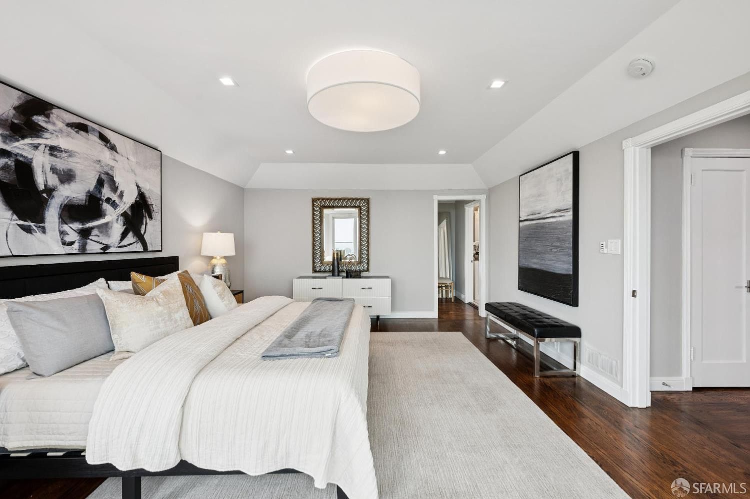 This is a primary bedroom featuring a large bed with a black headboard and white bedding, complemented by decorative pillows. The room is decorated with modern art pieces and a dresser with a mirror. The flooring is dark hardwood, and a light-colored rug covers a large portion of the floor, creating a cozy and inviting atmosphere.