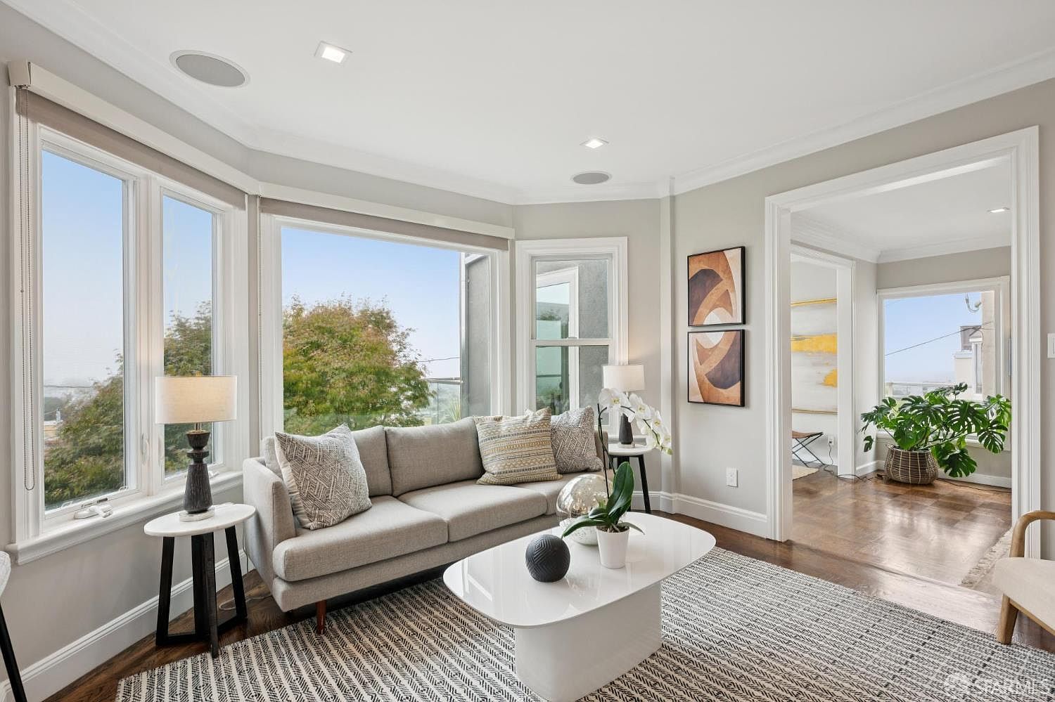 This is a bright and airy living room featuring a neutral color palette with a gray sofa, a white coffee table, and a patterned rug. Large windows offer a view of the outdoors, and artwork adorns the walls. The space is well-lit with recessed lighting and lamps, creating a comfortable and inviting atmosphere.