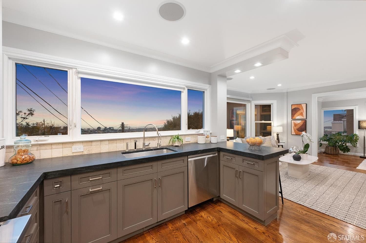 This is a well-lit kitchen featuring gray cabinetry, dark countertops, and stainless steel appliances. A large window above the sink offers a scenic view of the outside. The hardwood flooring adds warmth to the space, which seamlessly transitions into an adjacent living area.