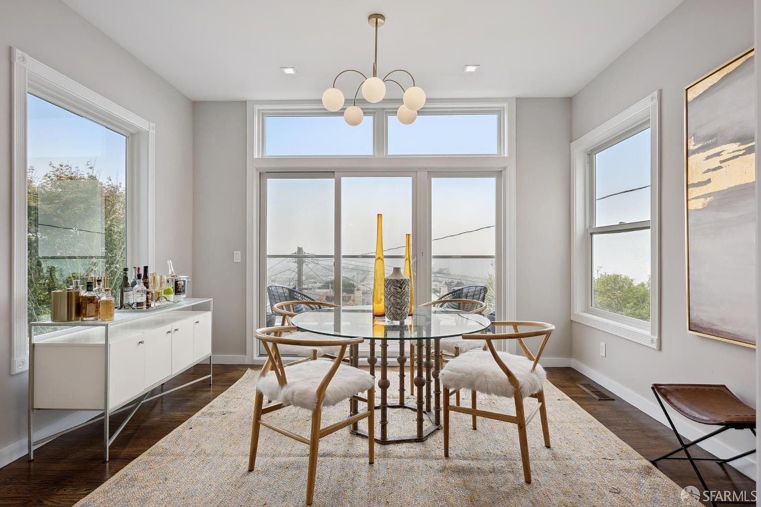 This is a bright and airy dining room featuring a round glass table with unique metal base, surrounded by four wooden chairs with white faux fur cushions. A modern chandelier hangs above the table, and a large window offers a scenic view. A white sideboard with a mirrored top sits against the wall, adding to the room's elegance.