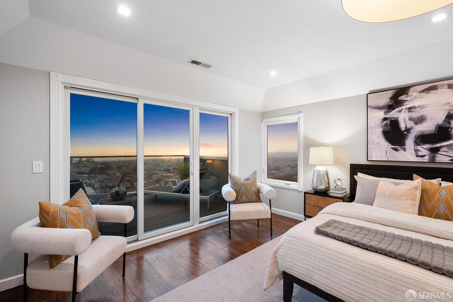 This is a primary bedroom featuring a modern design with hardwood floors and a neutral color palette. The room includes a king-size bed with decorative pillows, two armchairs, and a sliding glass door leading to a balcony with city views. An abstract art piece hangs above the bed, adding a touch of sophistication.