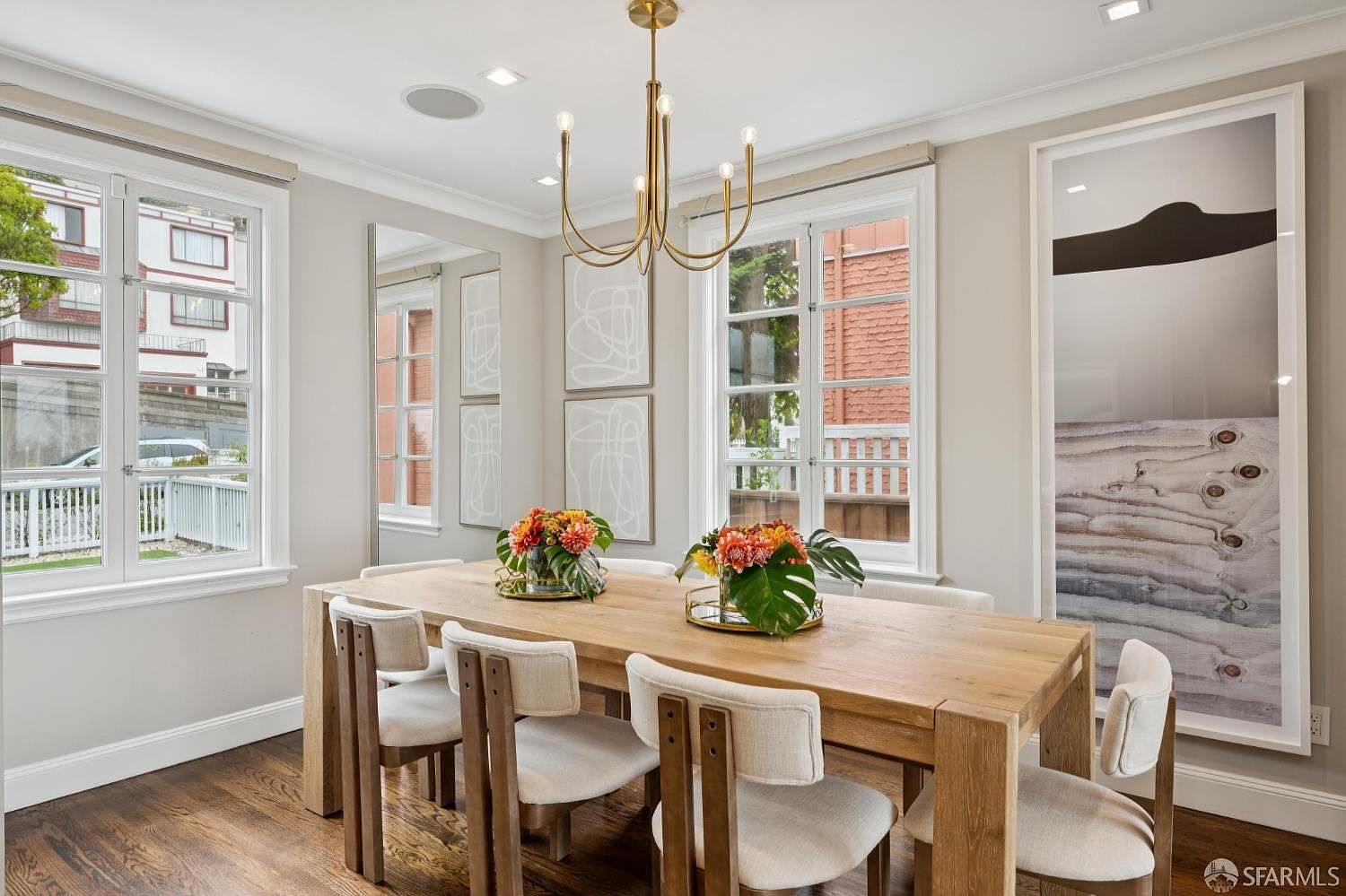 This is an interior shot of a dining room featuring a large wooden table with seating for eight. The room is well-lit with natural light from the windows and an elegant chandelier. Artwork adorns the walls, and floral arrangements serve as centerpieces, creating a warm and inviting atmosphere.