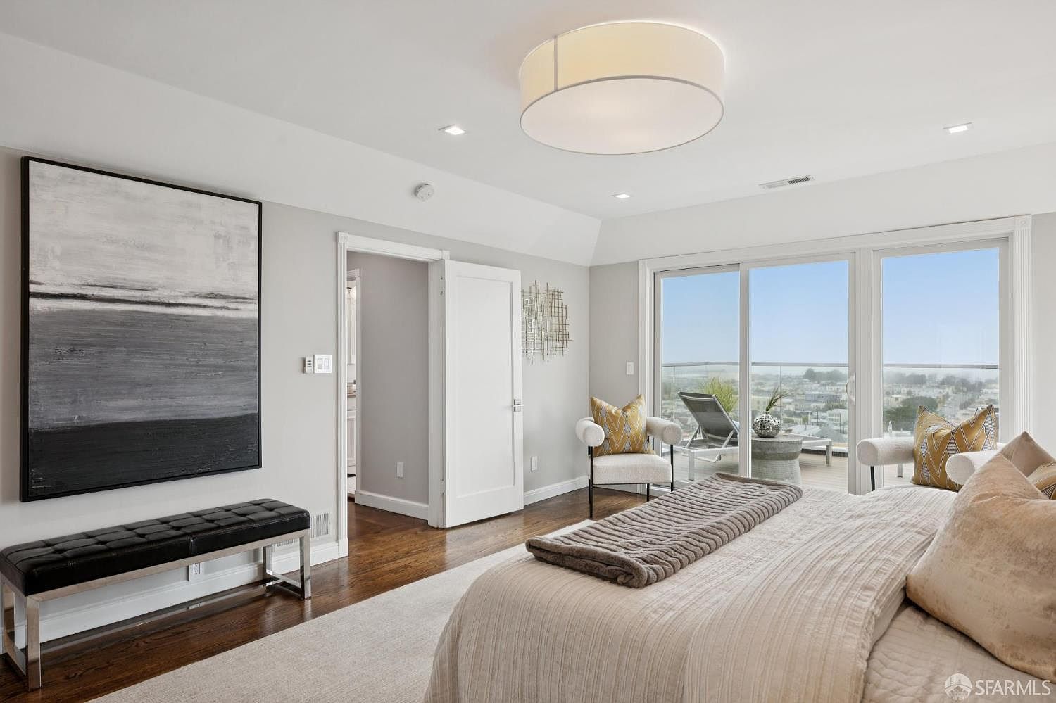 This is a bright and spacious primary bedroom featuring hardwood floors, a large abstract painting, and a comfortable-looking bed with neutral-toned bedding. Sliding glass doors lead to a balcony with city views, enhancing the room's appeal. The room is decorated in a modern style with a minimalist aesthetic.