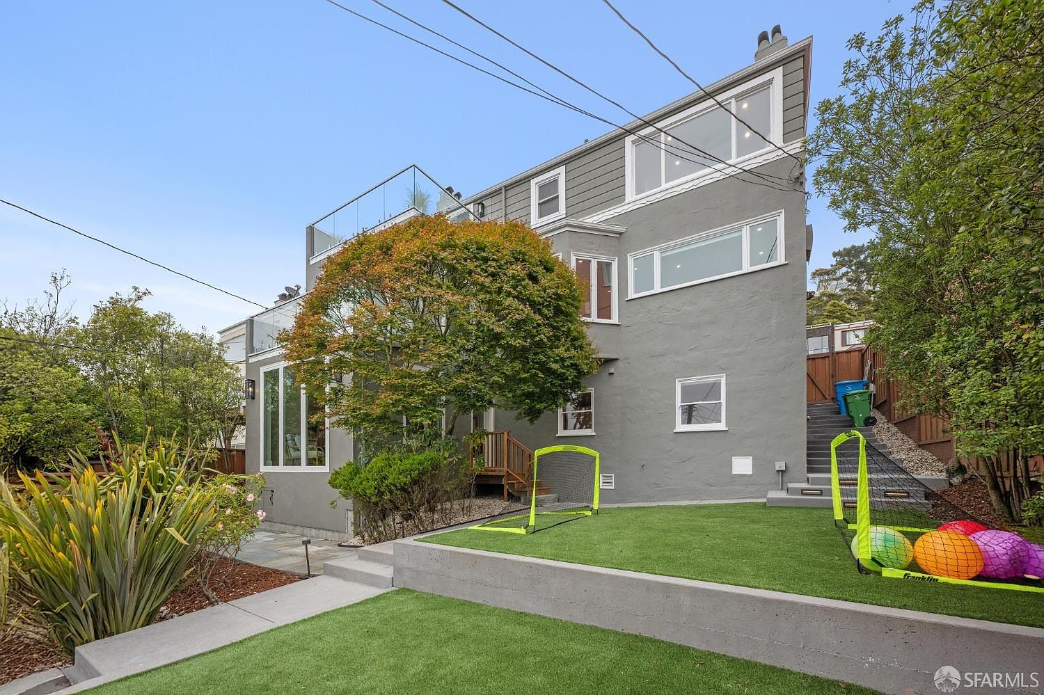 This is a rear view of a multi-story gray house with a modern design. The backyard features a well-maintained lawn with artificial turf, a concrete retaining wall, and a small soccer goal. A wooden staircase leads up to a higher level of the property, and mature trees and landscaping add to the privacy and appeal of the outdoor space.