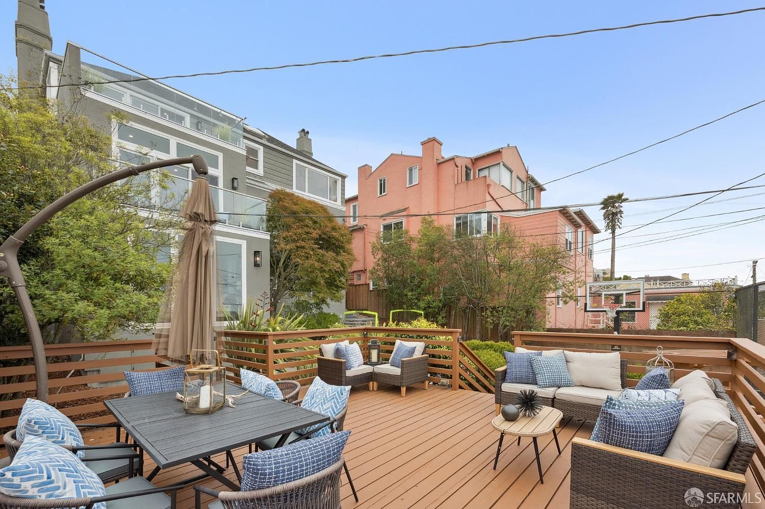 This image showcases a well-appointed outdoor deck area, featuring comfortable seating arrangements with plush cushions and a dining table set for entertaining. The deck is constructed with wood and surrounded by a wooden railing, offering a sense of privacy and enclosure. The backdrop includes residential buildings and greenery, creating a pleasant outdoor living space.