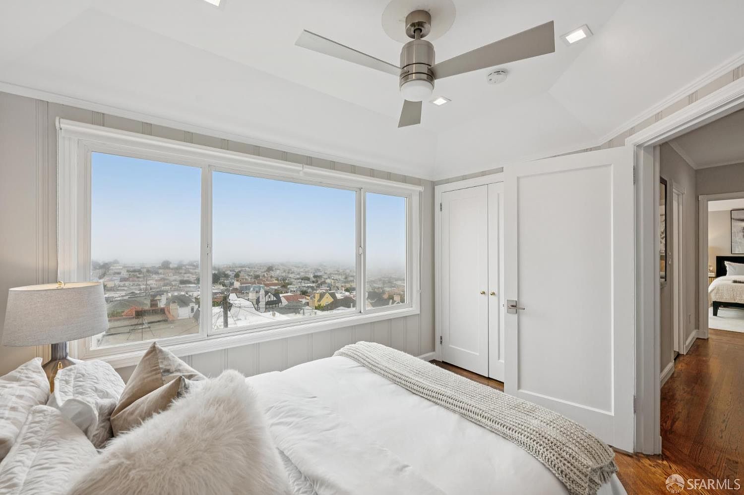 This is a bright and airy primary bedroom featuring a large window with a city view. The room is decorated in neutral tones, with white walls, bedding, and light wood floors. A ceiling fan provides air circulation, and there is a closet and doorway visible, suggesting ample storage and access to other areas of the home.
