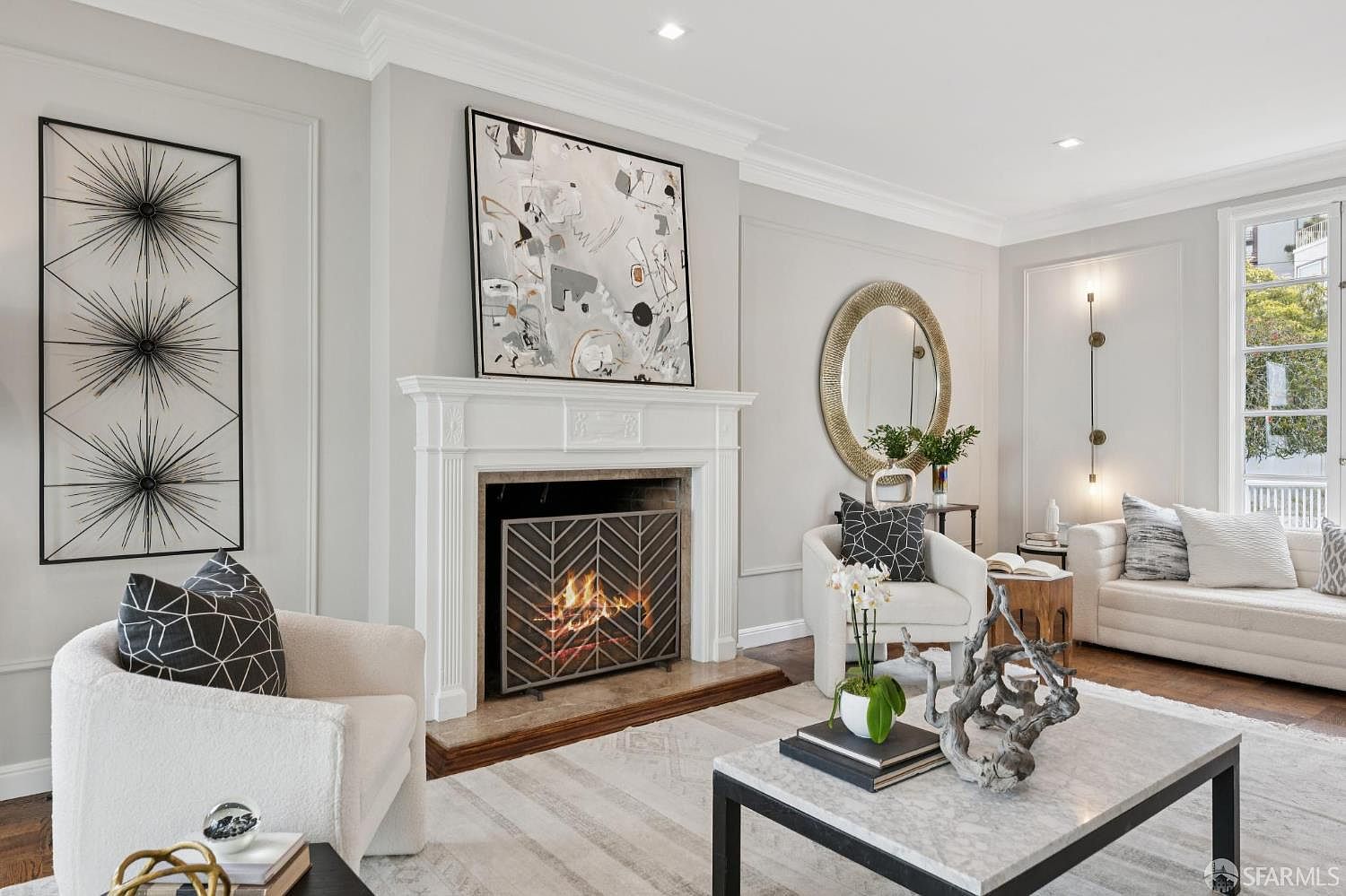 This is an interior shot of a living room featuring a fireplace with a decorative screen and an abstract painting above it. The room is decorated in neutral tones with white armchairs, a marble-topped coffee table, and modern art pieces on the wall. The overall impression is elegant and contemporary.