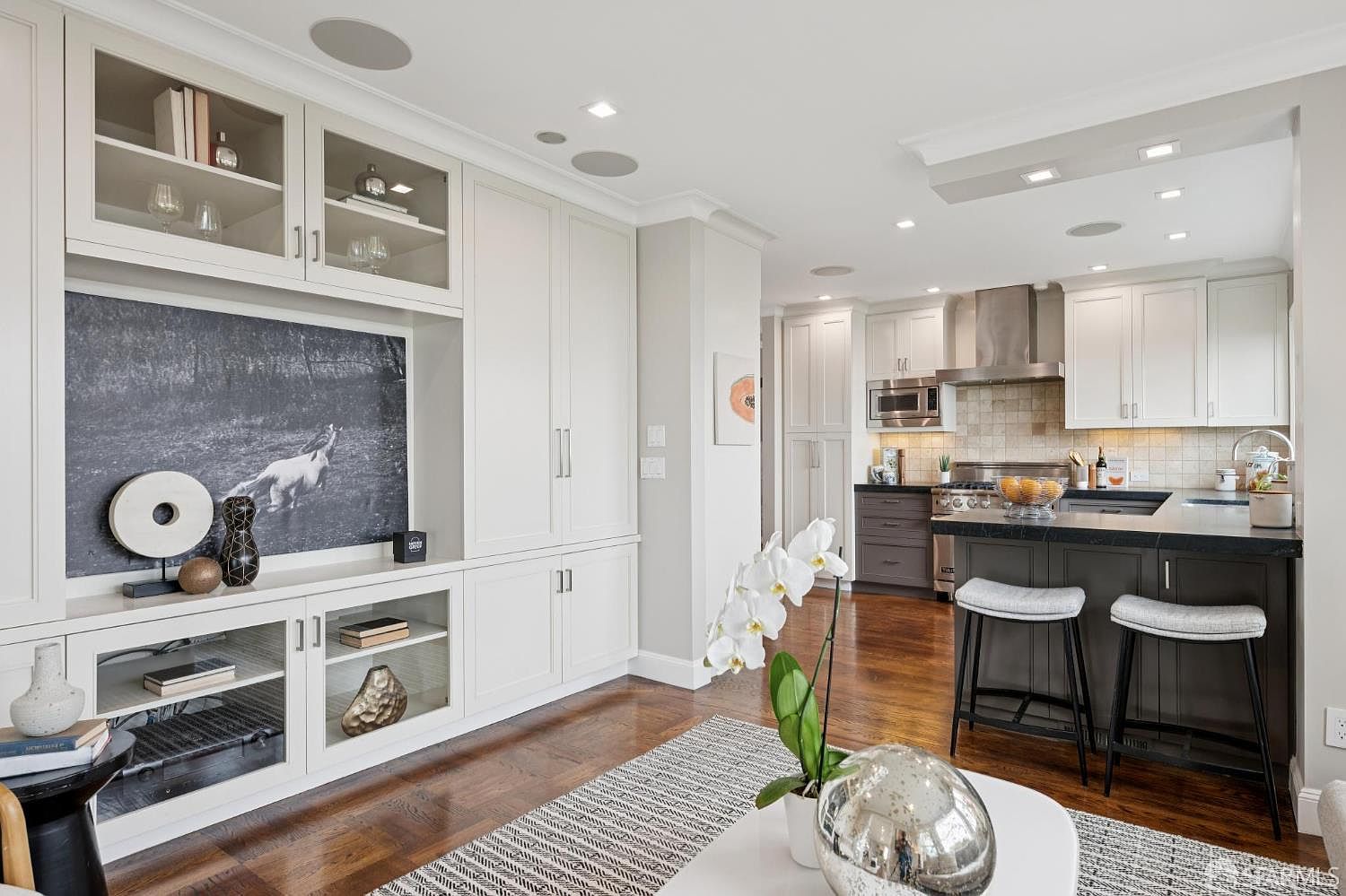 This image showcases a stylish living room that seamlessly transitions into a modern kitchen. The living area features custom built-in cabinetry with display shelves and a large black and white art piece, while the kitchen boasts stainless steel appliances, dark countertops, and bar stool seating. The hardwood floors and neutral color palette create a sophisticated and inviting atmosphere.