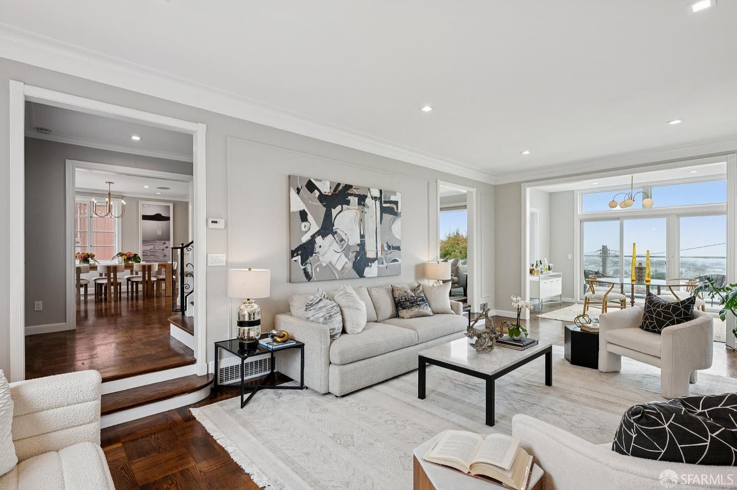 This is an interior shot of a well-lit living room featuring a neutral-toned sofa with decorative pillows, a marble-topped coffee table, and an abstract art piece on the wall. The room opens to a dining area and offers a glimpse of an outdoor view through large windows, creating a bright and inviting atmosphere. The flooring is a mix of hardwood and a light-colored area rug, adding texture and warmth to the space.