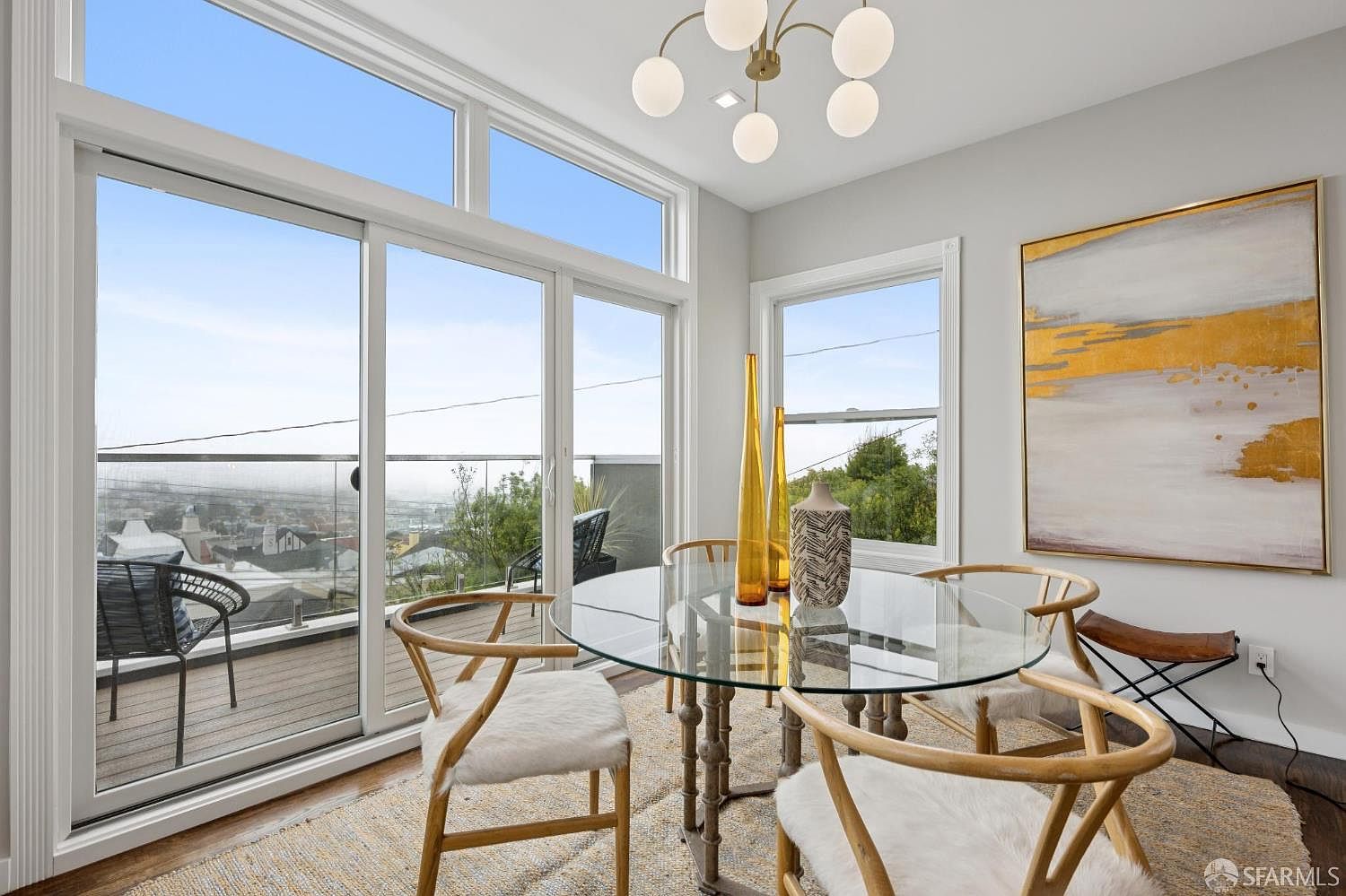 This is a bright and airy dining room featuring a round glass table with unique wooden chairs accented with white fur. The room has large windows offering a scenic view, complemented by a modern light fixture and an abstract painting on the wall. The space is well-lit and stylish, creating an inviting atmosphere for dining.