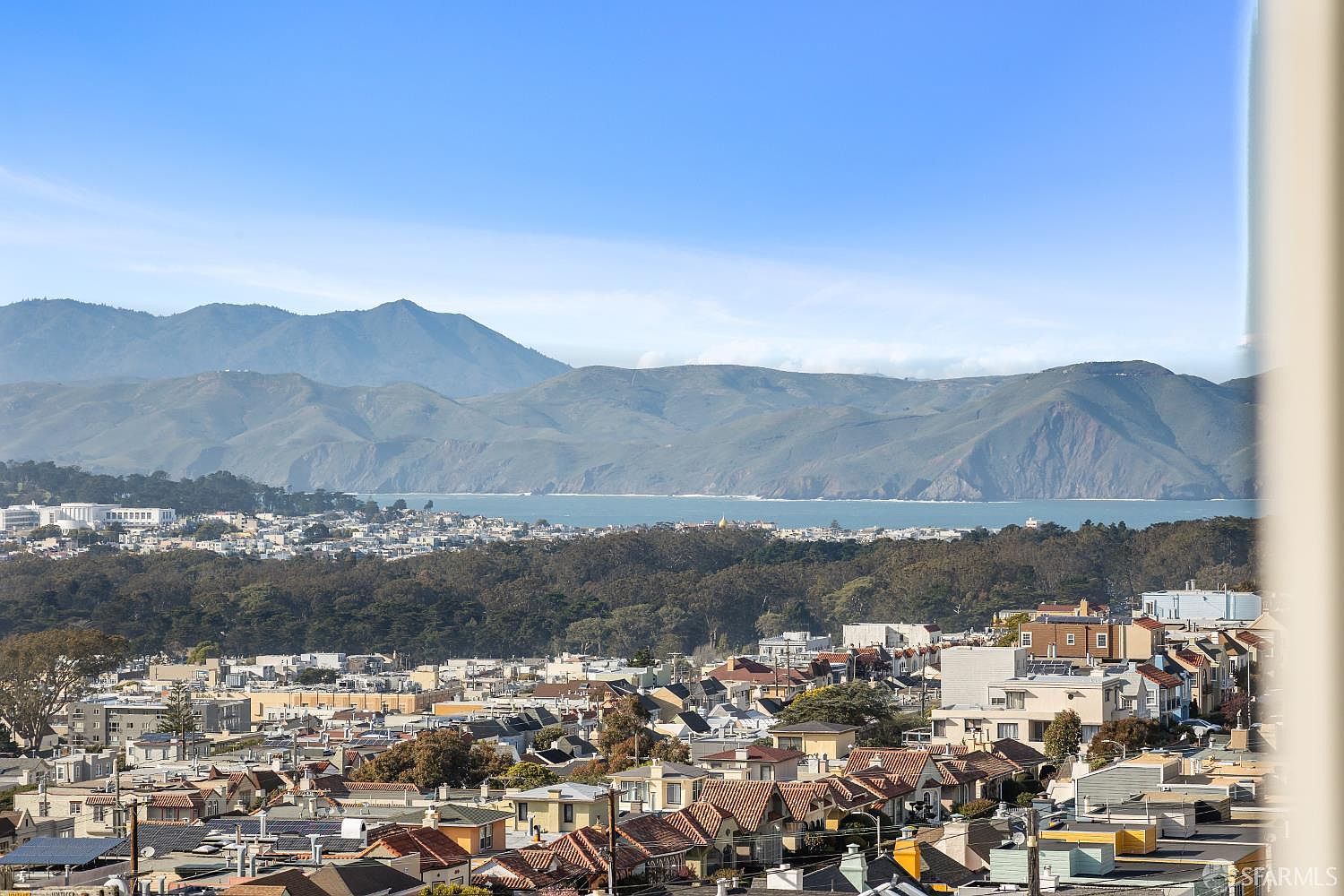 This aerial view showcases a dense residential neighborhood with a mix of building styles and red-tiled roofs. In the background, rolling hills and a body of water create a scenic backdrop under a clear blue sky. The image provides a sense of the neighborhood's layout and its proximity to natural landscapes.