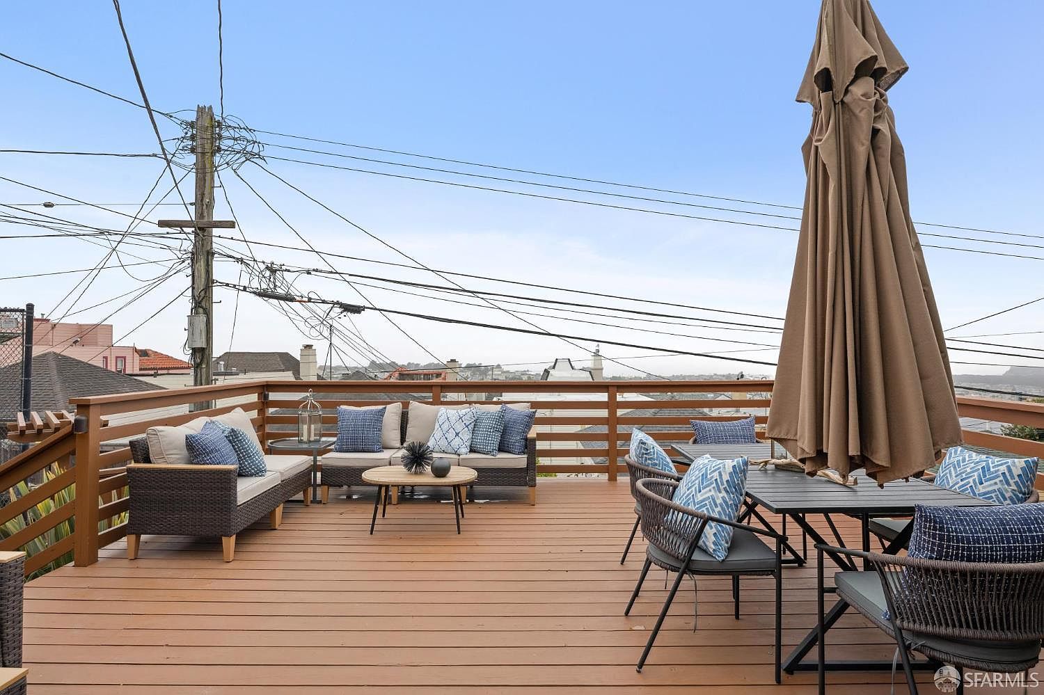 This image showcases a well-furnished outdoor deck area, perfect for relaxation and entertainment. The deck features comfortable seating arrangements, including a sofa and chairs with stylish blue and white cushions, arranged around a coffee table and dining table. A large, closed umbrella provides shade, and the wooden deck offers a warm, inviting atmosphere with a view of the surrounding neighborhood.