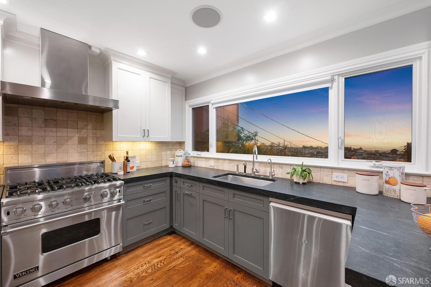 This is a well-lit kitchen featuring stainless steel appliances, including a Viking range and a dishwasher. The cabinetry is a mix of white upper cabinets and gray lower cabinets, complemented by dark countertops and a light tile backsplash. A large window provides natural light and a view of the outside, enhancing the kitchen's appeal.