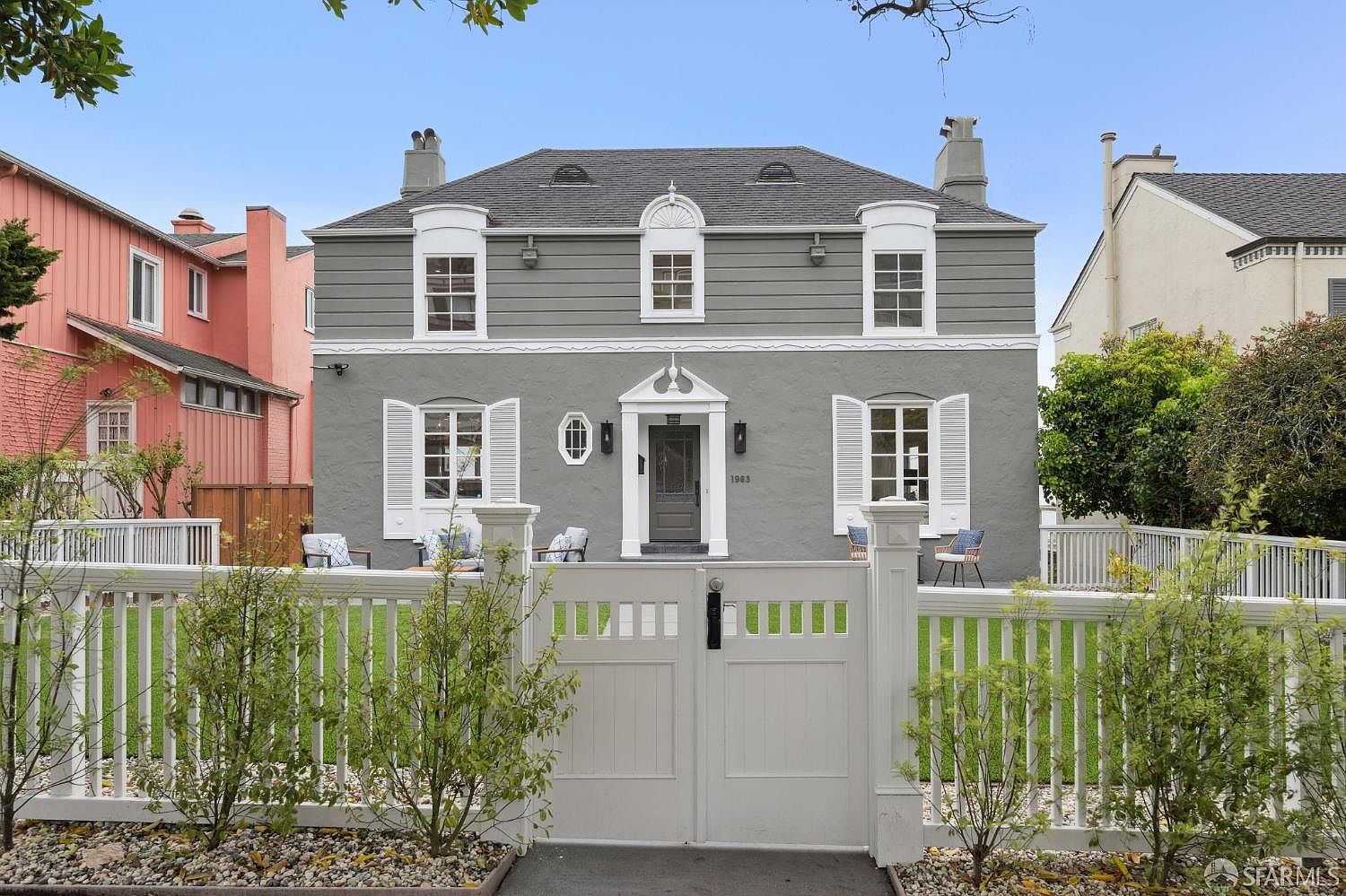 This is a charming front view of a two-story house with a gray exterior and white trim. The house features symmetrical windows with shutters, a central front door with decorative accents, and a well-maintained front yard with a white picket fence and gate. The overall impression is one of classic elegance and curb appeal.