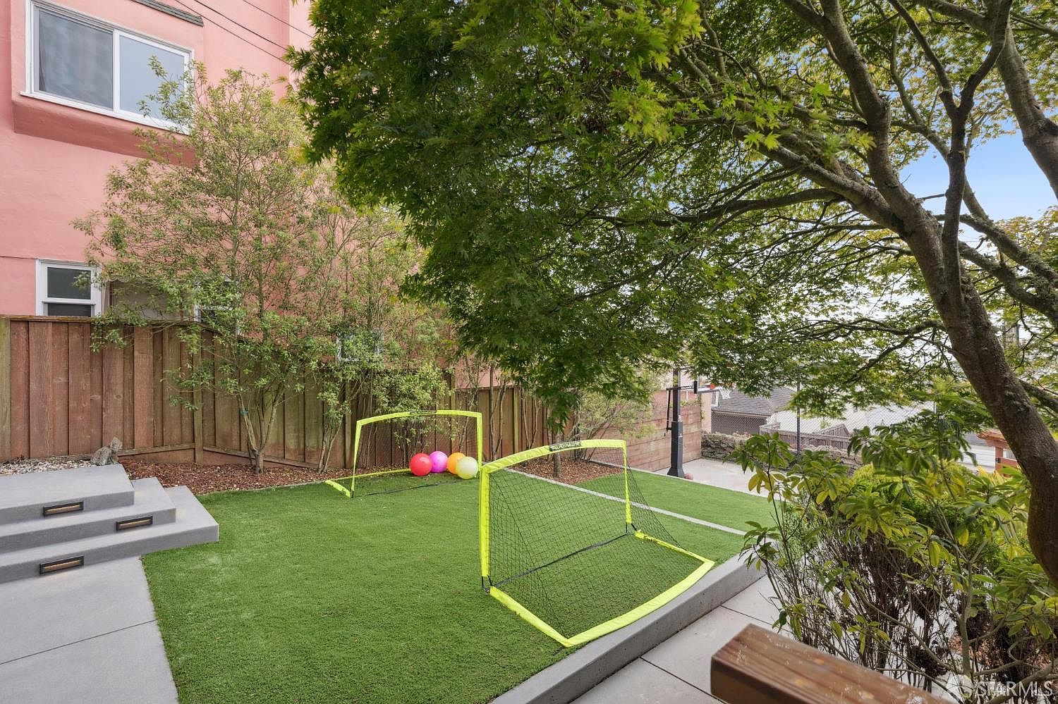 This image showcases a well-maintained backyard featuring artificial turf, two small soccer goals, and a set of concrete steps leading to a higher level. The yard is bordered by a wooden fence and lush greenery, including trees and shrubs. The perspective is from a slightly elevated position, offering a comprehensive view of the outdoor space, suggesting a private and recreational area.