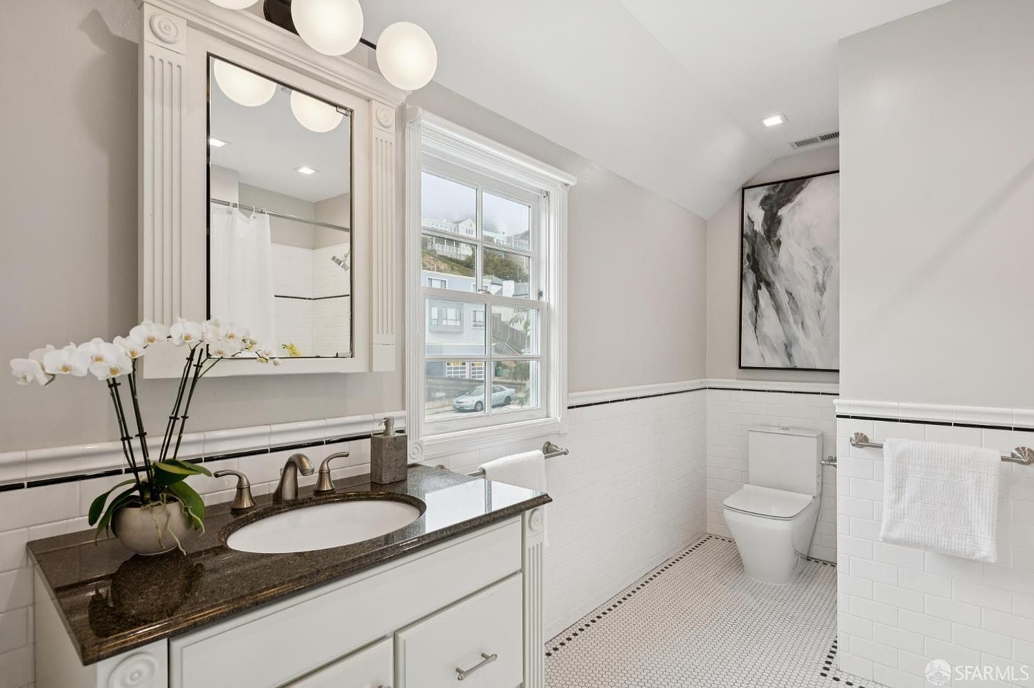 This is a well-lit bathroom featuring a white vanity with a dark countertop, a decorative mirror, and a window providing natural light. The walls are partially tiled with white subway tiles accented by a black stripe, and the floor is covered in small white hexagonal tiles with a black border. A toilet and an abstract art piece are visible in the background, contributing to the bathroom's clean and classic aesthetic.