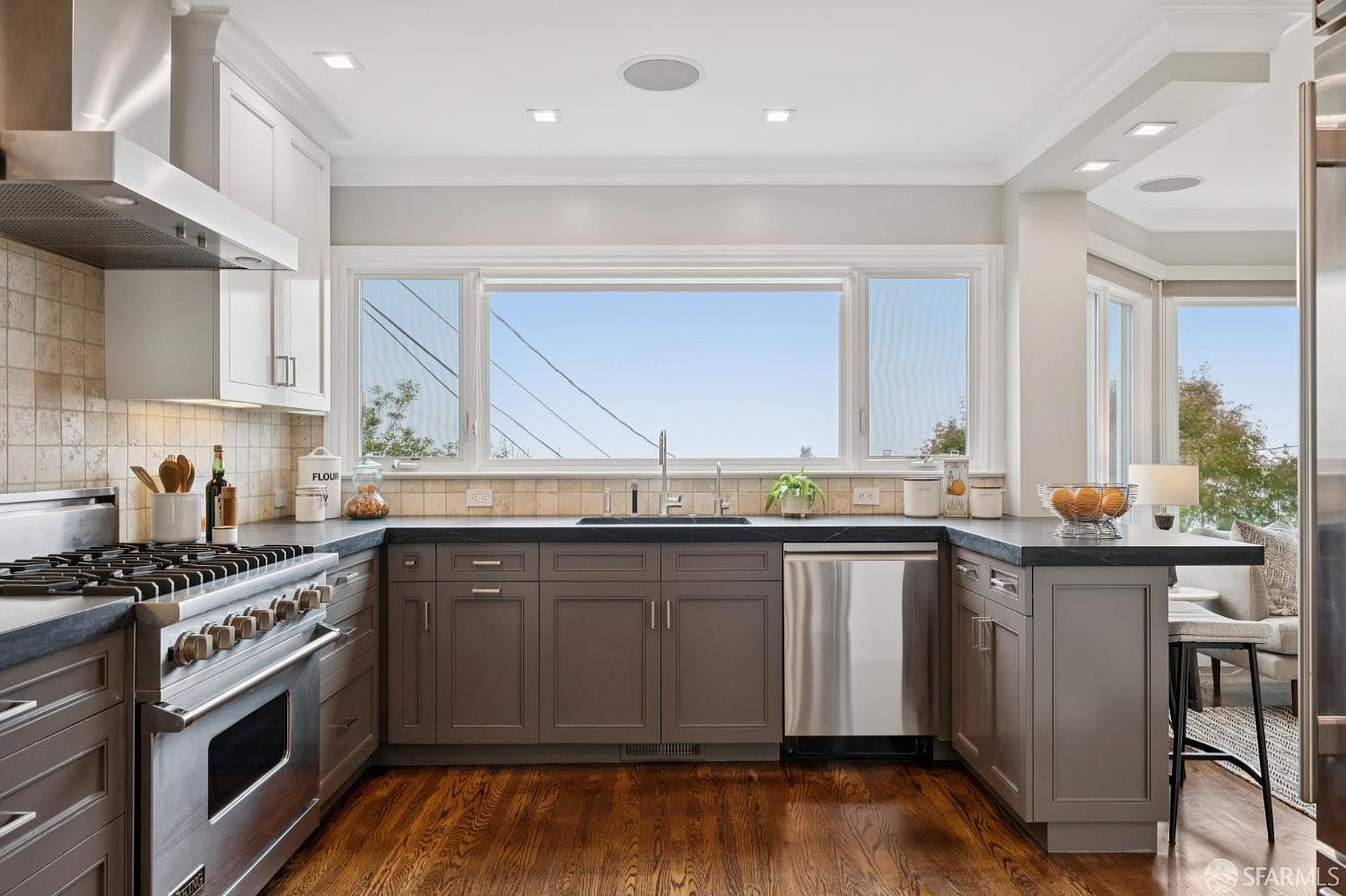 This is a well-lit kitchen featuring dark countertops and a large window providing ample natural light. The kitchen is equipped with stainless steel appliances and a gas range. The hardwood flooring adds warmth to the space, creating an inviting atmosphere.