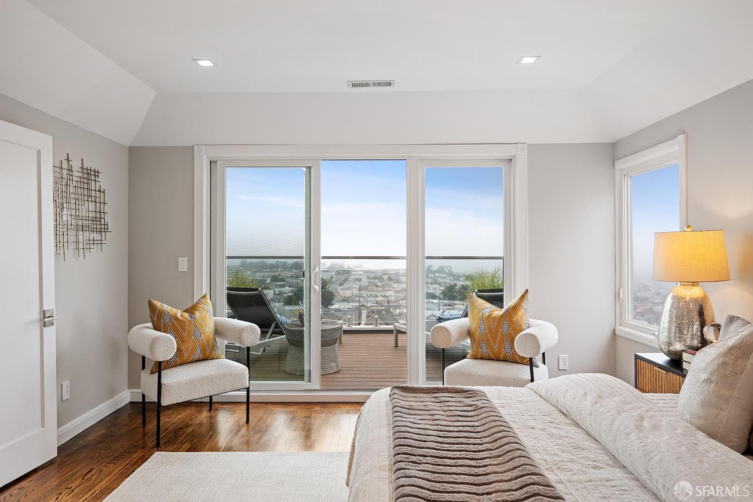 This is a bright and airy primary bedroom featuring a sliding glass door that leads to a balcony with city views. The room is furnished with two armchairs, a bed with neutral-toned bedding, and a bedside table with a lamp. The hardwood floors and neutral wall colors create a serene and inviting atmosphere.