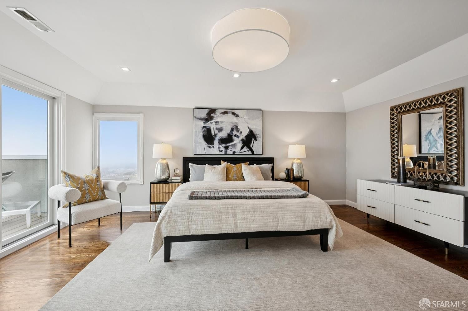 This is a well-lit primary bedroom featuring a large bed with a black frame, flanked by matching nightstands and lamps. An abstract art piece hangs above the bed, and a decorative mirror sits atop a white dresser on the right. The room is spacious, with hardwood floors partially covered by a large area rug, and a chair sits near a window offering a view.