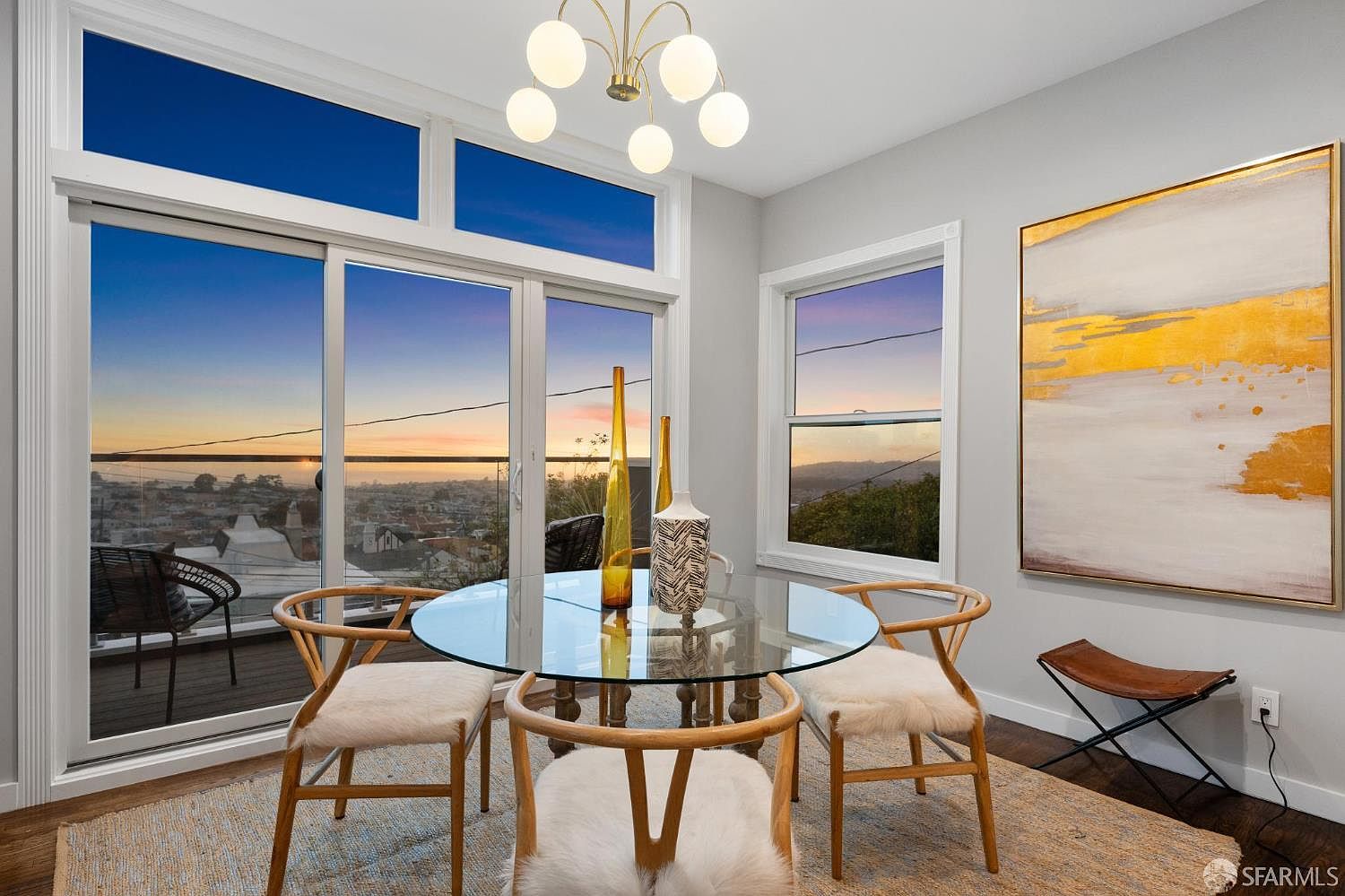This dining room features a round glass table surrounded by four chairs with fur seat covers. Large windows offer a stunning view of the city at sunset, complemented by a modern chandelier. An abstract painting hangs on the wall, adding a touch of elegance to the space.
