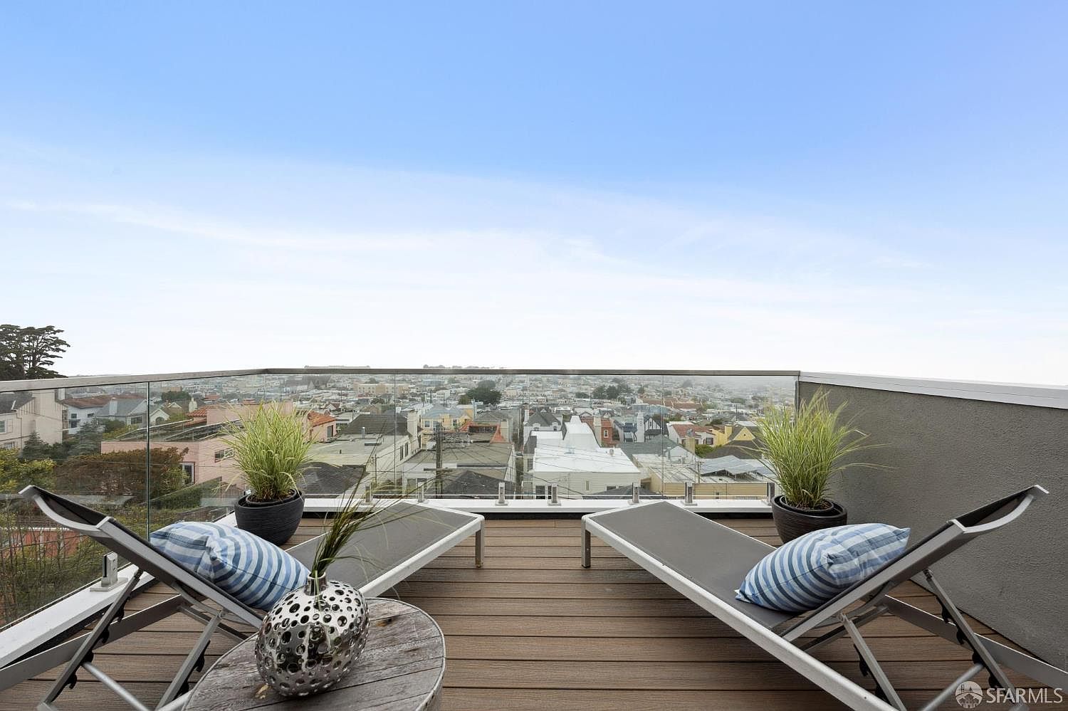 This image showcases a stylish rooftop deck with panoramic city views. The deck features modern lounge chairs with striped pillows, a small decorative table, and potted plants, creating a relaxing outdoor space. A glass railing provides an unobstructed view of the cityscape, enhancing the property's appeal.