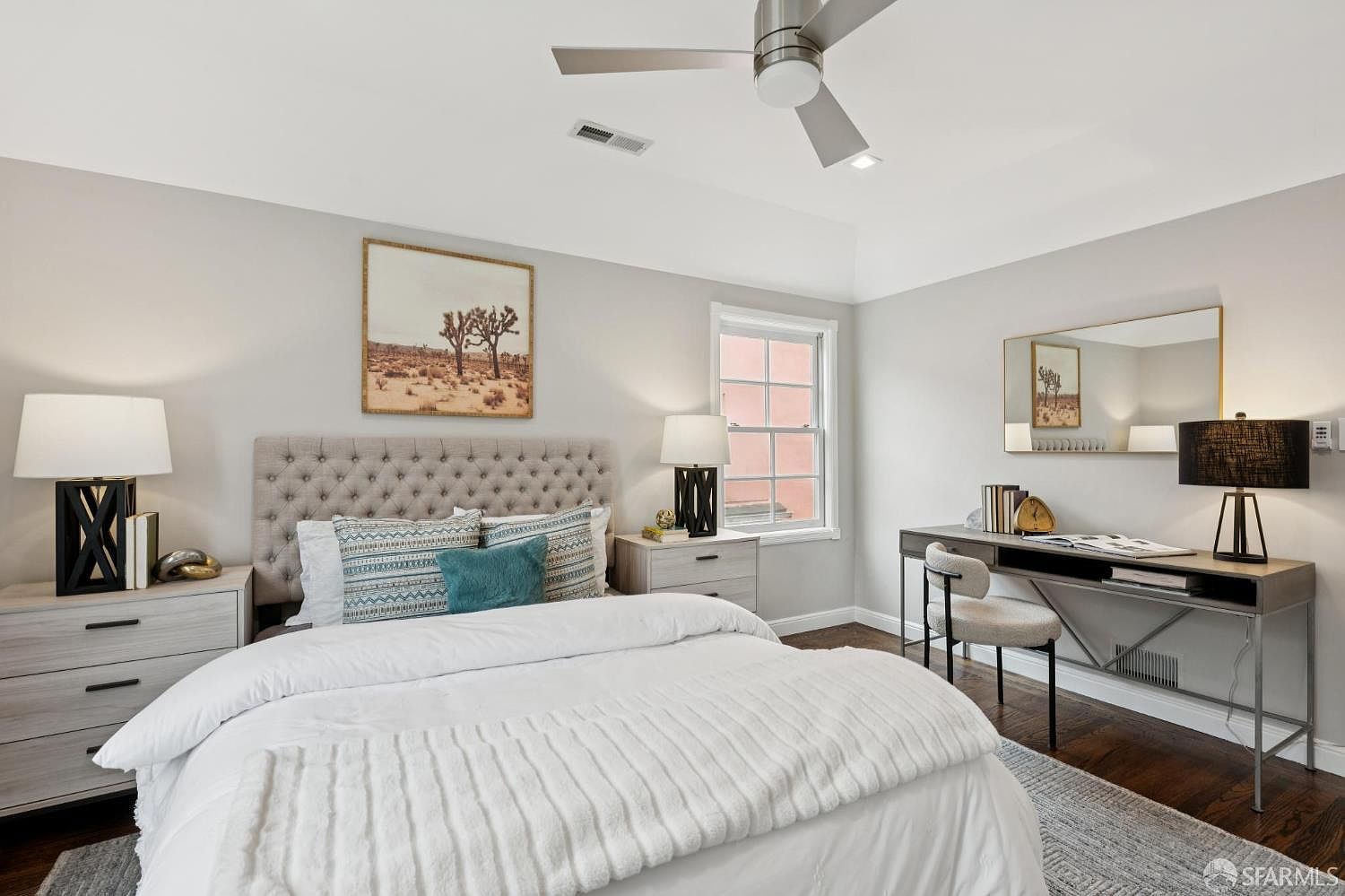 This is a well-lit primary bedroom featuring a tufted headboard, a white duvet, and a neutral color palette. The room includes a desk area with a mirror, a nightstand with a lamp, and artwork depicting a desert landscape. The overall impression is calm and inviting, with a focus on comfort and style.
