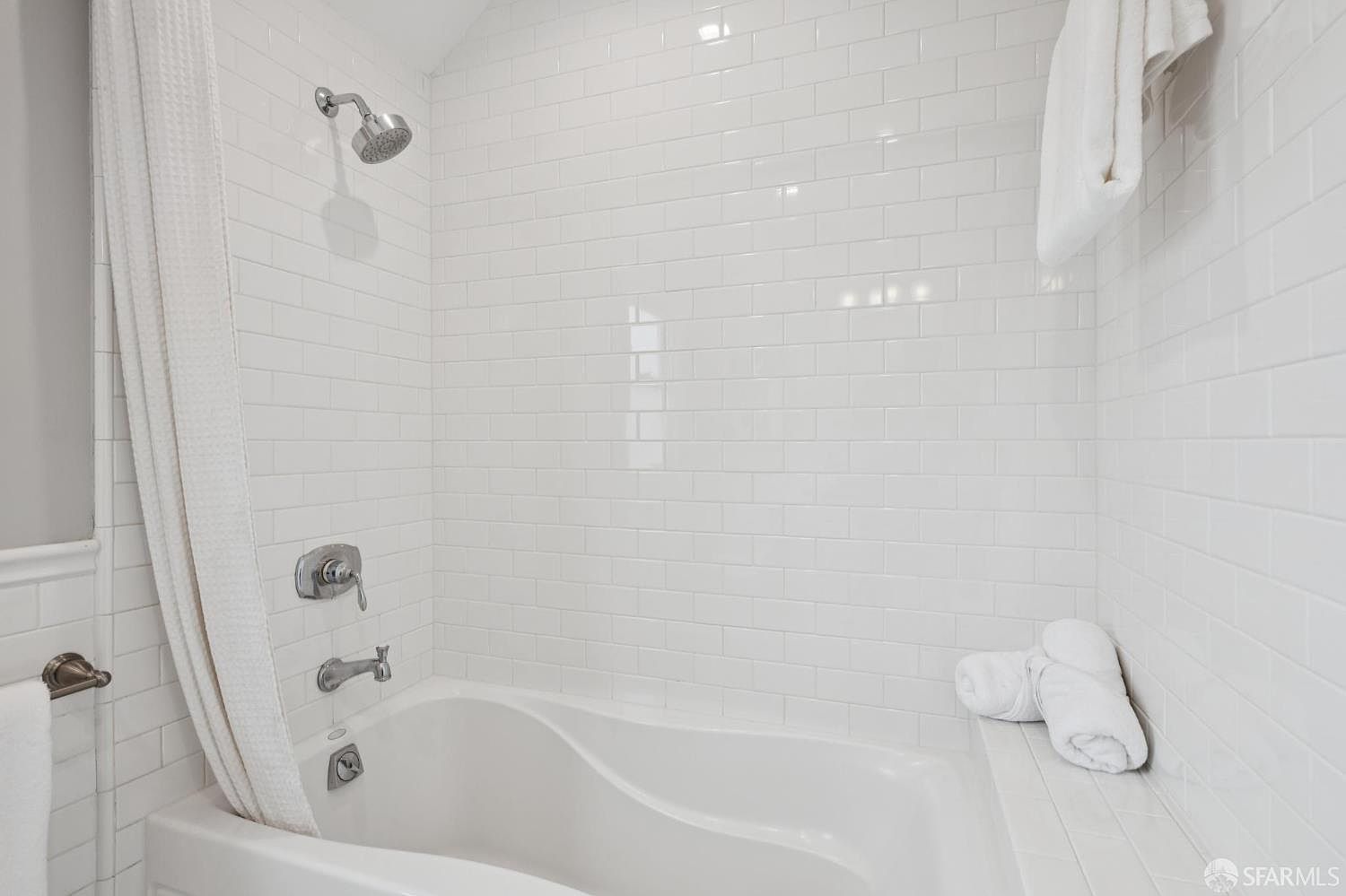 This is a well-lit bathroom featuring a white subway tile surround and a white bathtub. A shower curtain is partially drawn, and there are neatly folded white towels on the ledge of the tub. The overall impression is clean and modern.