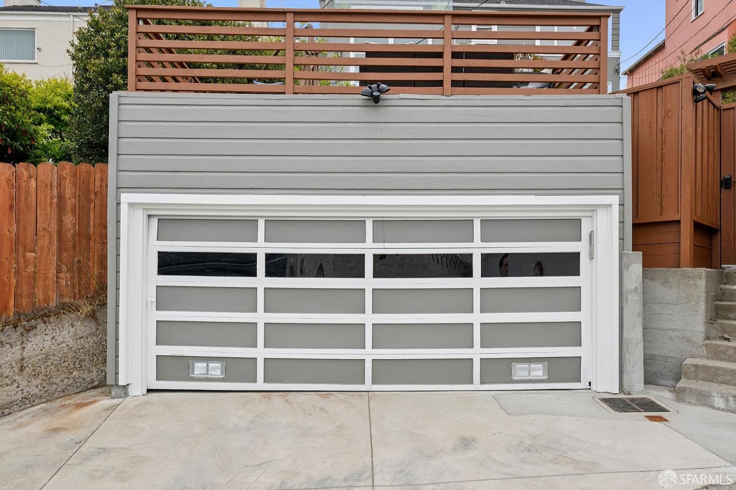 The image showcases a modern garage exterior featuring a gray and white garage door with multiple rectangular windows. The garage is painted gray with horizontal siding, and a wooden fence is visible on the side. The concrete driveway leads up to the garage, and a wooden deck with a railing is situated above the garage structure.