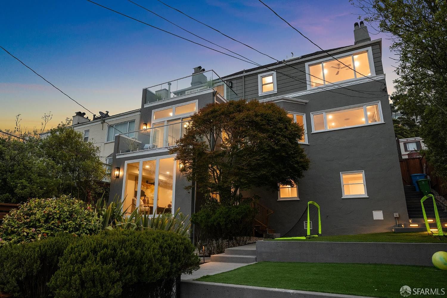 The image showcases the rear exterior of a multi-story home with a modern design. Large windows and glass-railed balconies offer ample natural light and outdoor living space. The property features a well-maintained lawn and landscaping, enhancing its curb appeal.
