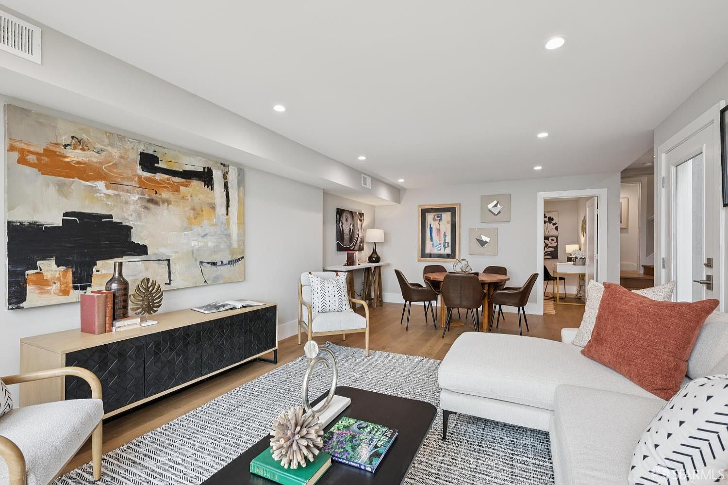 This is an interior shot of a living room featuring a large abstract painting above a modern cabinet. The room includes a seating area with a sectional sofa, a coffee table with books, and a dining area with a round table and chairs. The space is well-lit with recessed lighting and natural light, creating a welcoming and stylish atmosphere.