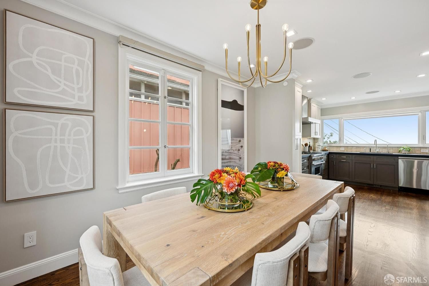 This is an interior shot of a dining room featuring a large wooden table with white upholstered chairs. The room is decorated with modern artwork and a stylish chandelier. The dining area flows into a kitchen space, creating an open and inviting atmosphere.