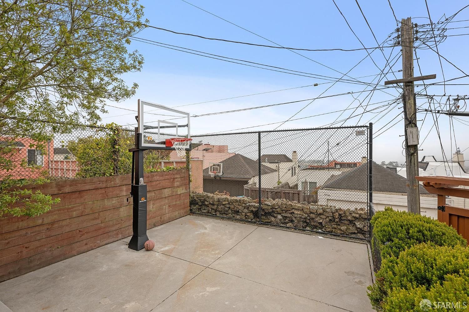 This image showcases a compact outdoor basketball court area. The court is enclosed by a wooden fence on one side and a chain-link fence with a stone base on the other, providing privacy and containment. A portable basketball hoop stands ready for play, and the concrete surface is clean and well-maintained, suggesting a functional and enjoyable recreational space.