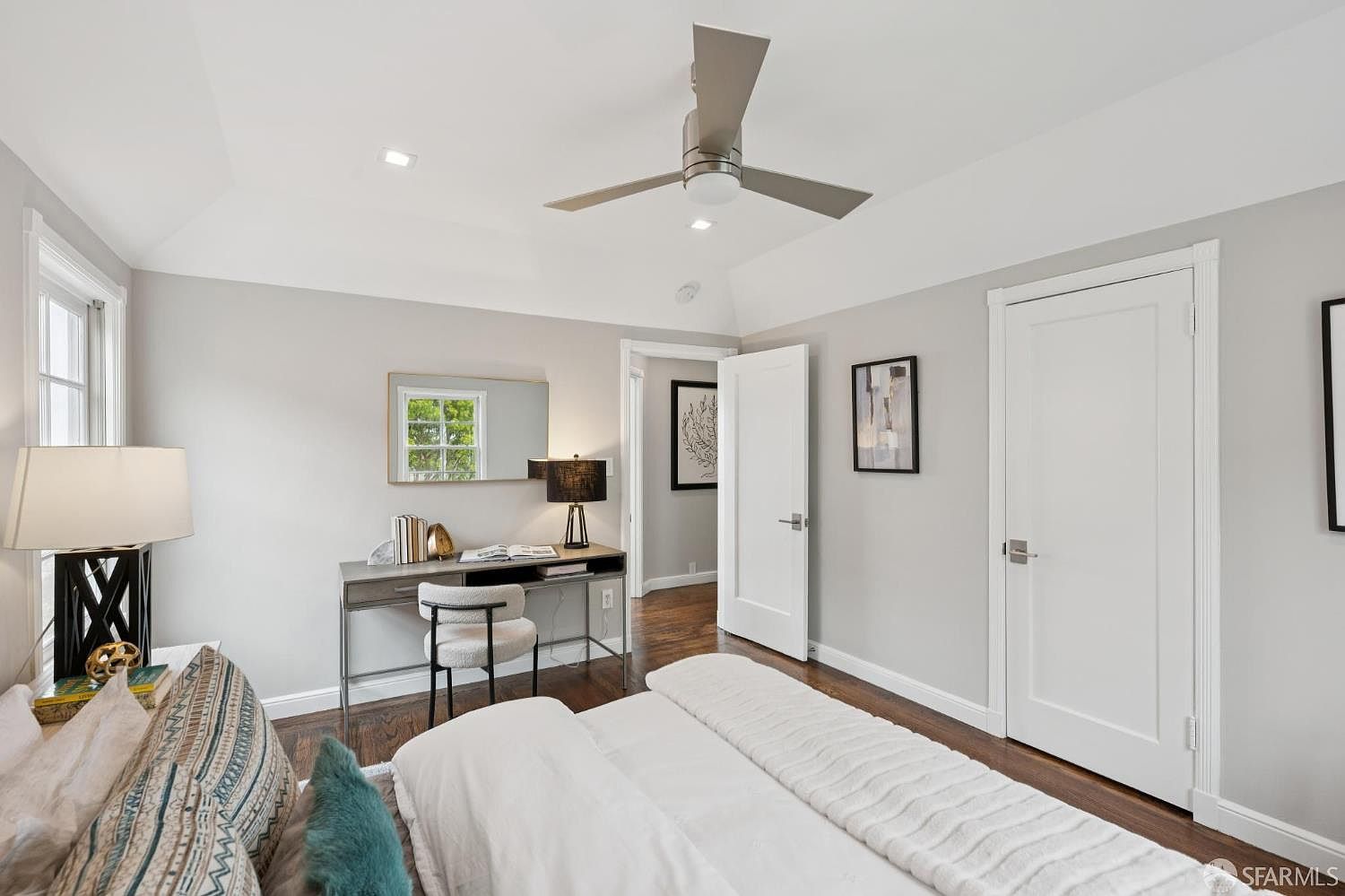 This is a bright and airy primary bedroom featuring a neutral color palette with hardwood floors. The room includes a desk area with a mirror and a chair, as well as a ceiling fan. The bed is neatly made with white linens and decorative pillows, creating a serene and inviting atmosphere.