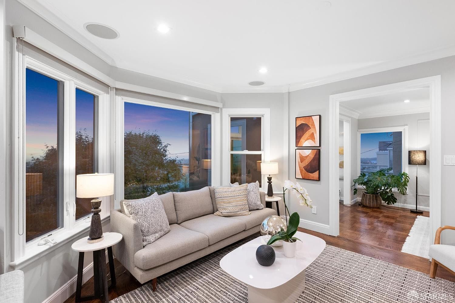 This is an interior shot of a living room featuring a neutral-toned sofa with decorative pillows, a white coffee table with decorative objects, and side tables with lamps. Large windows offer a view of the outside, and artwork adorns the walls. The room has a cozy and inviting atmosphere with a modern touch.